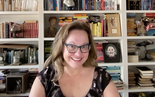Woman with glasses smiling in front of a bookshelf filled with books and decorative items.