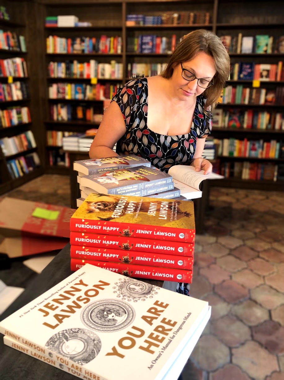 Woman reading a book at a table with stacks of books in a library, shelves filled with books in the background.