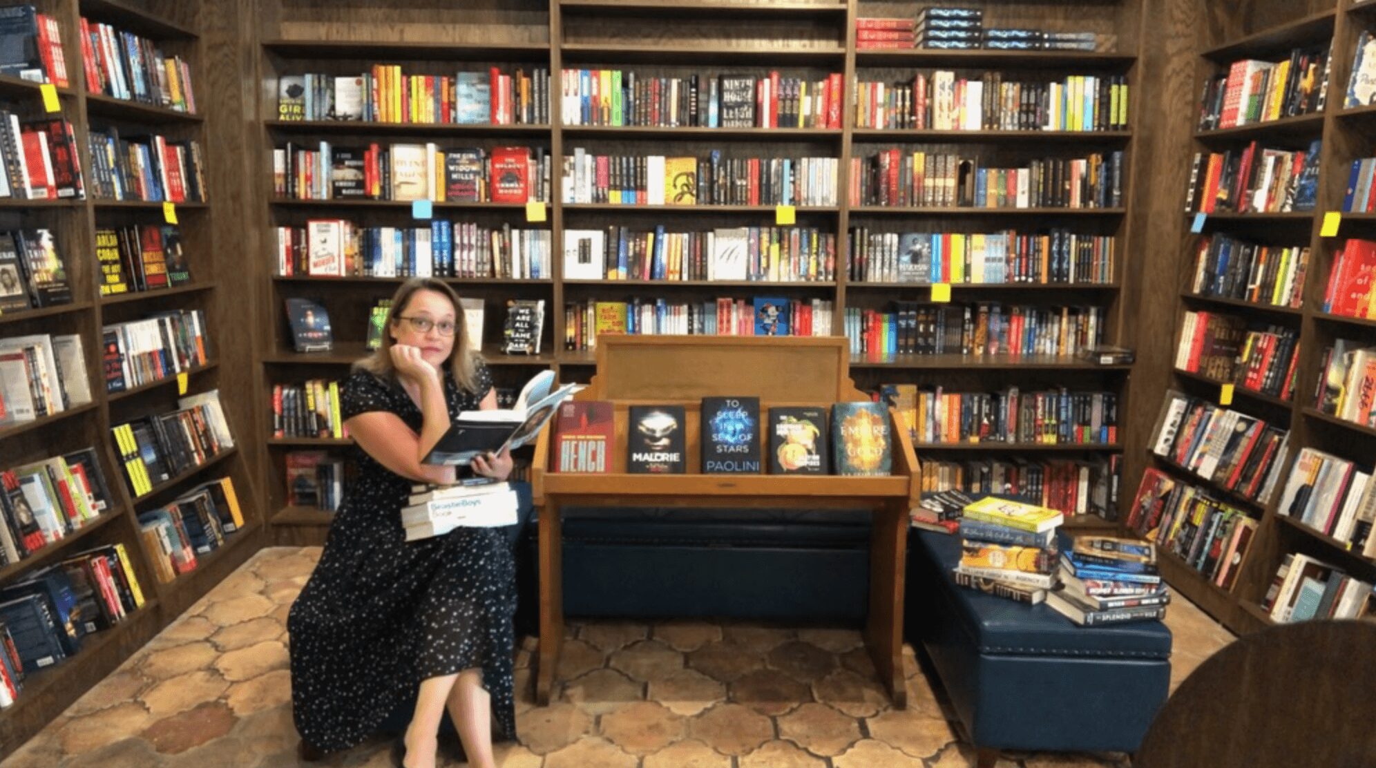 Woman sitting in a bookstore surrounded by tall bookshelves filled with books, holding a book and reading.