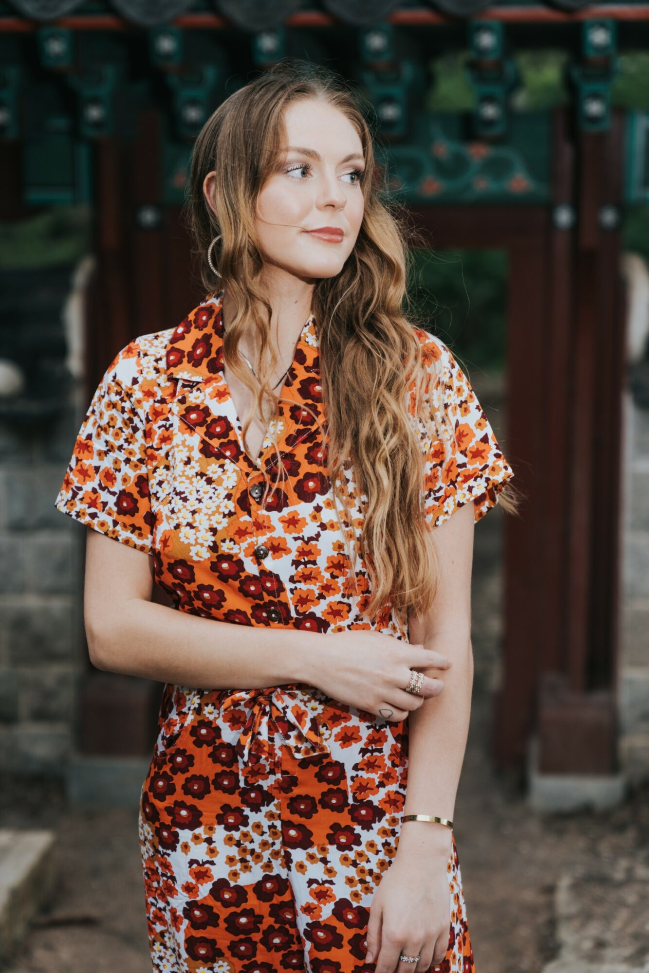Young woman with long wavy hair wearing a floral dress, standing outdoors in front of a wooden structure.