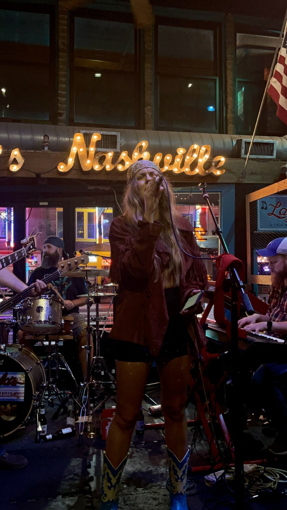 A woman singing into a microphone in a Nashville-themed bar with musicians and an American flag, illuminated by colorful lights.