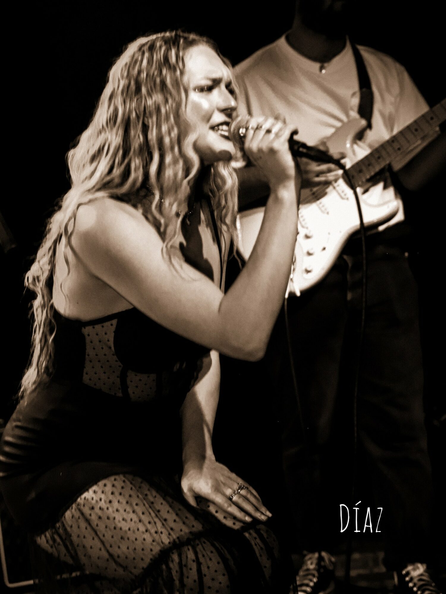 Woman singing into a microphone with a guitarist in the background, black and white image.