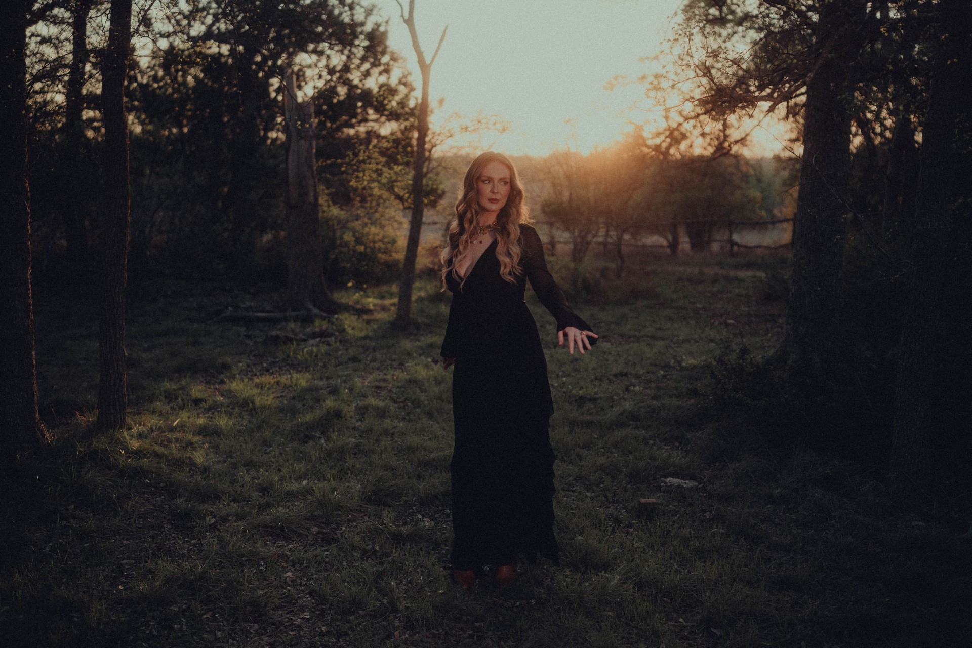 Woman standing outdoors in a forested area at sunset, wearing a long dark dress, with trees and sunlight in the background.