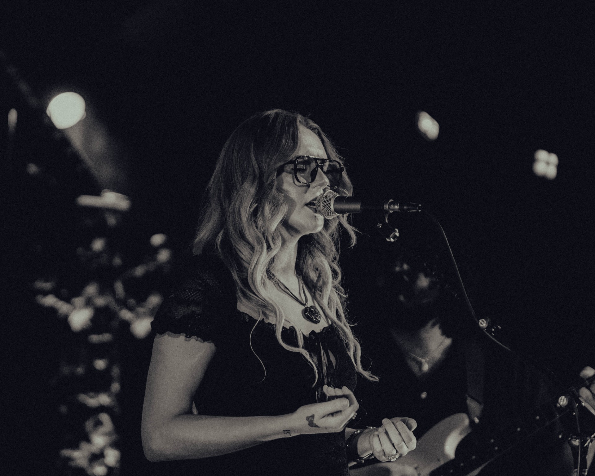 Woman with long wavy hair and glasses singing into a microphone on stage, with blurred lights in background.