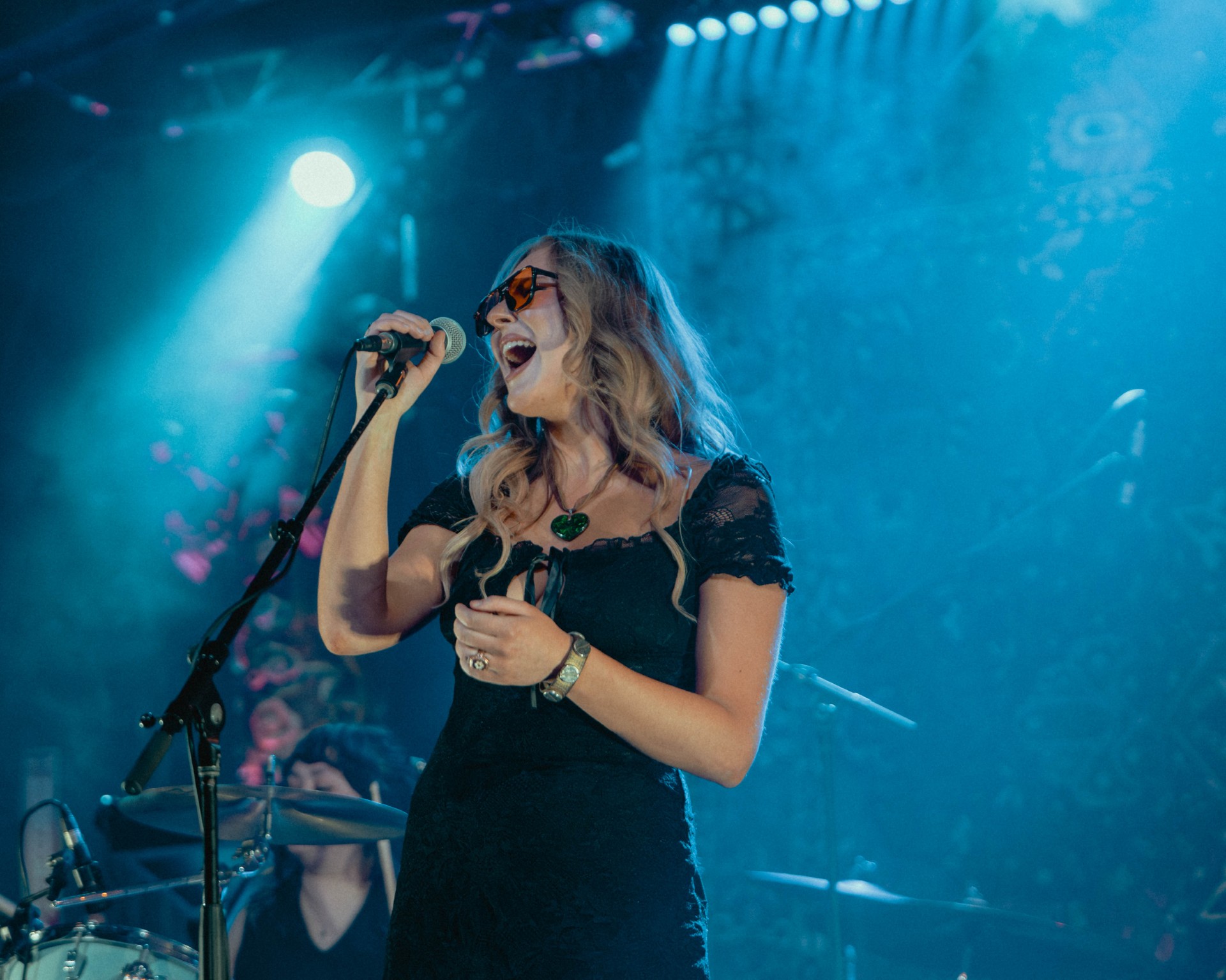 Woman singing into microphone on stage with blue lighting, wearing glasses and a black dress.