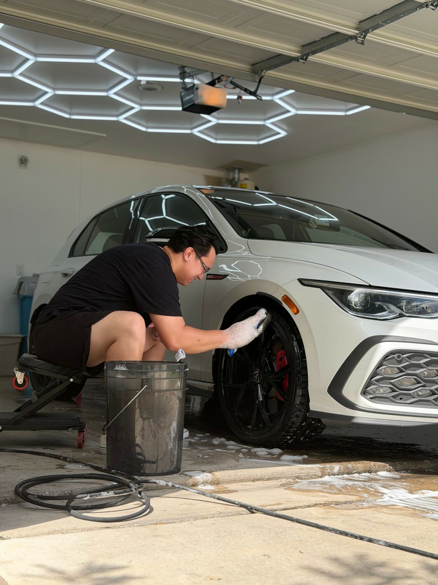 Person cleaning a white car's black wheel in a garage with hexagonal ceiling lights.