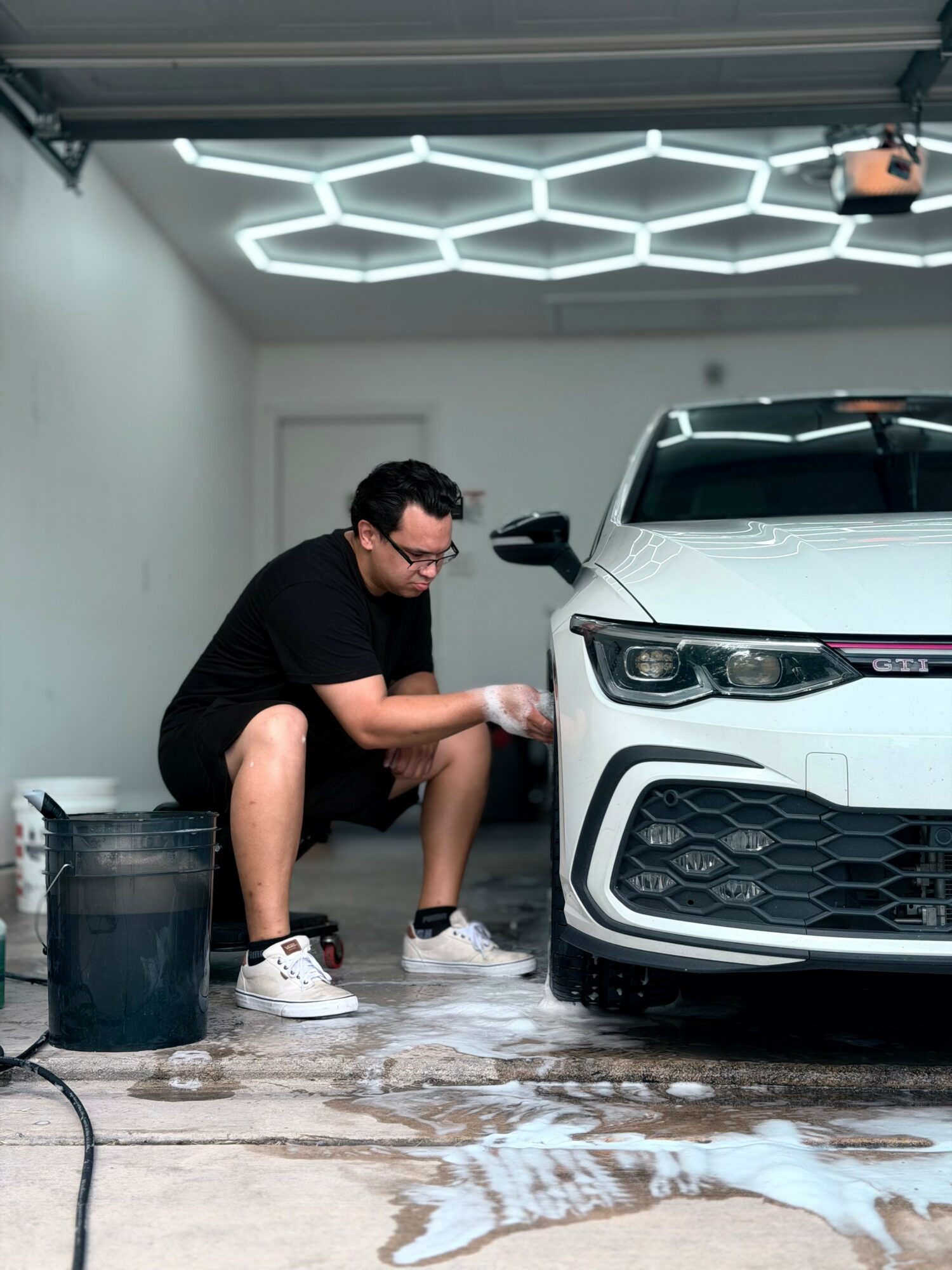 Person cleaning a white car inside a garage with hexagonal ceiling lights, wearing black shirt and shorts, with cleaning supplies nearby.