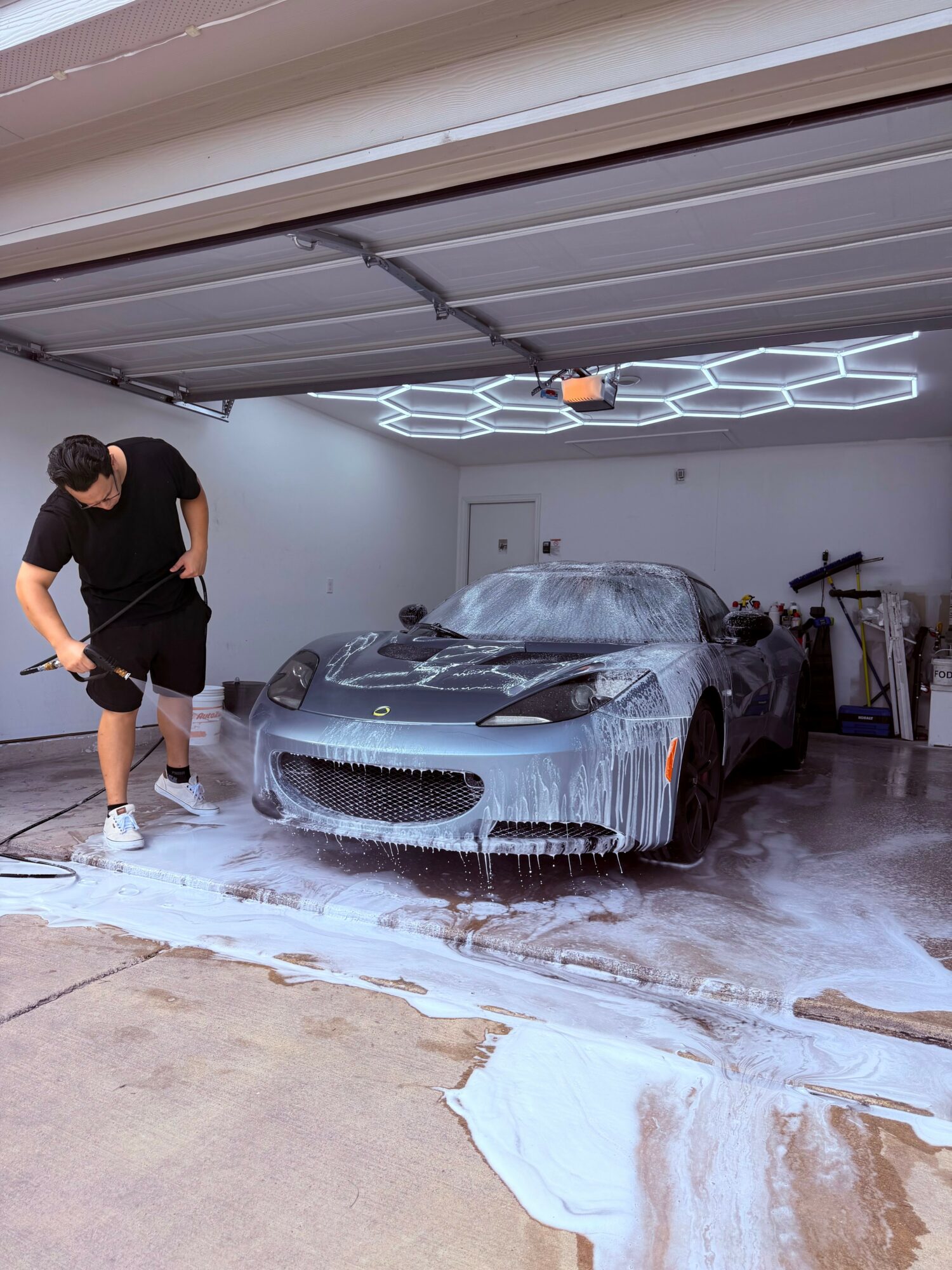 Person washing a sports car in a garage with foam and soap, garage door open, hexagonal light pattern on ceiling