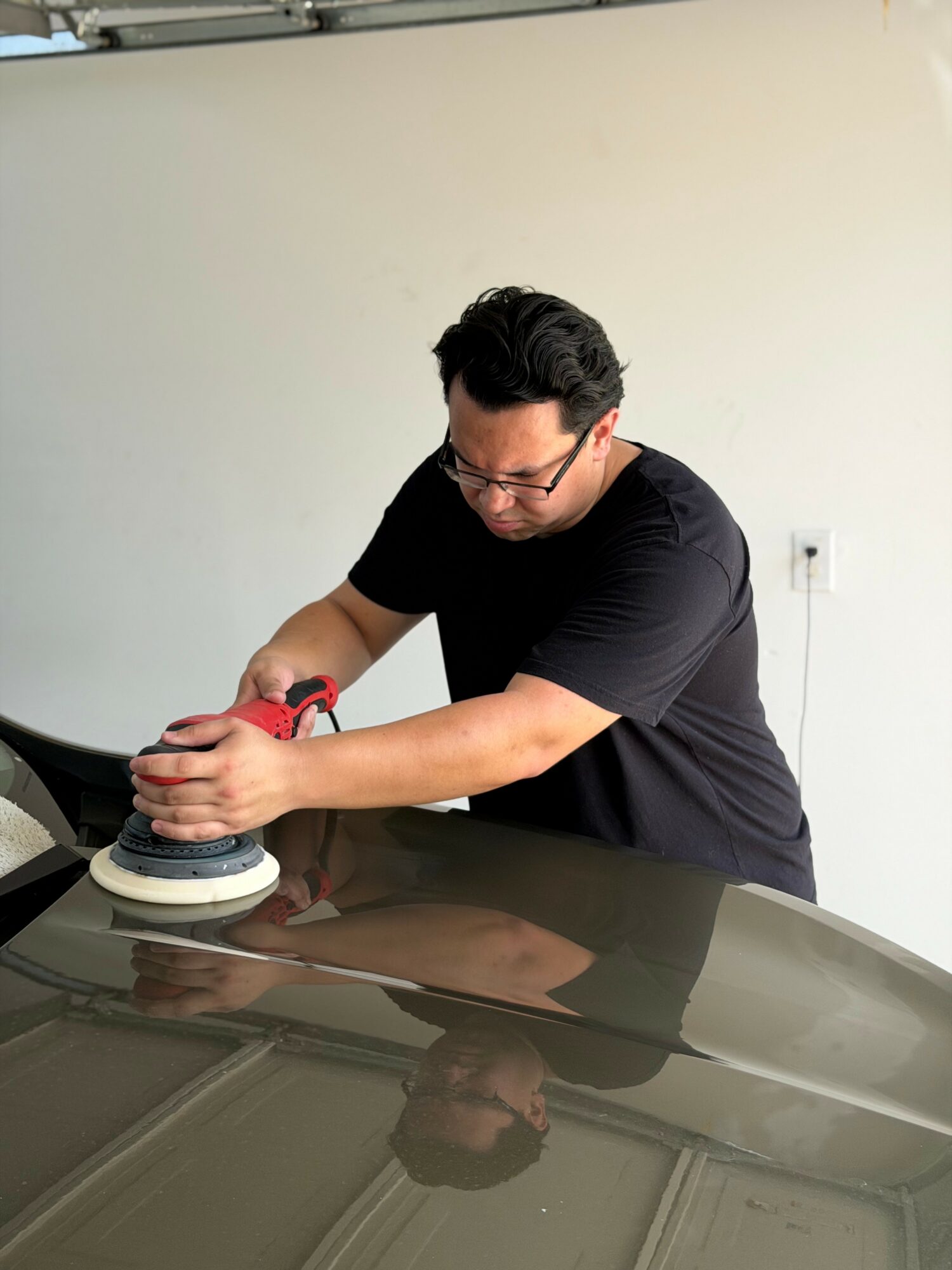 Person polishing a car with a handheld buffer, wearing glasses and a black shirt, in a plain room.