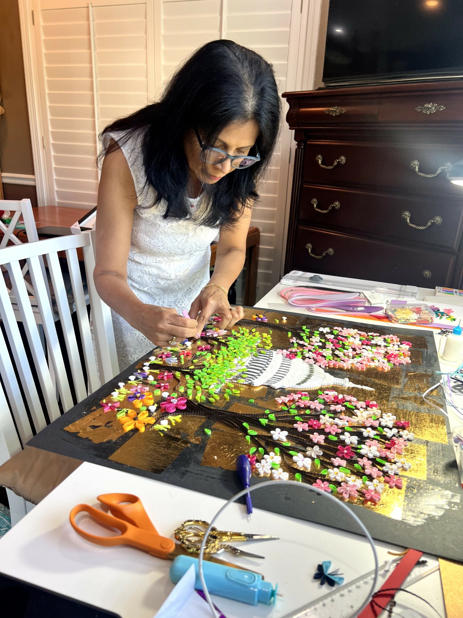 Woman arranging colorful artificial flowers on a table with scissors and craft supplies, in a room with wooden furniture.