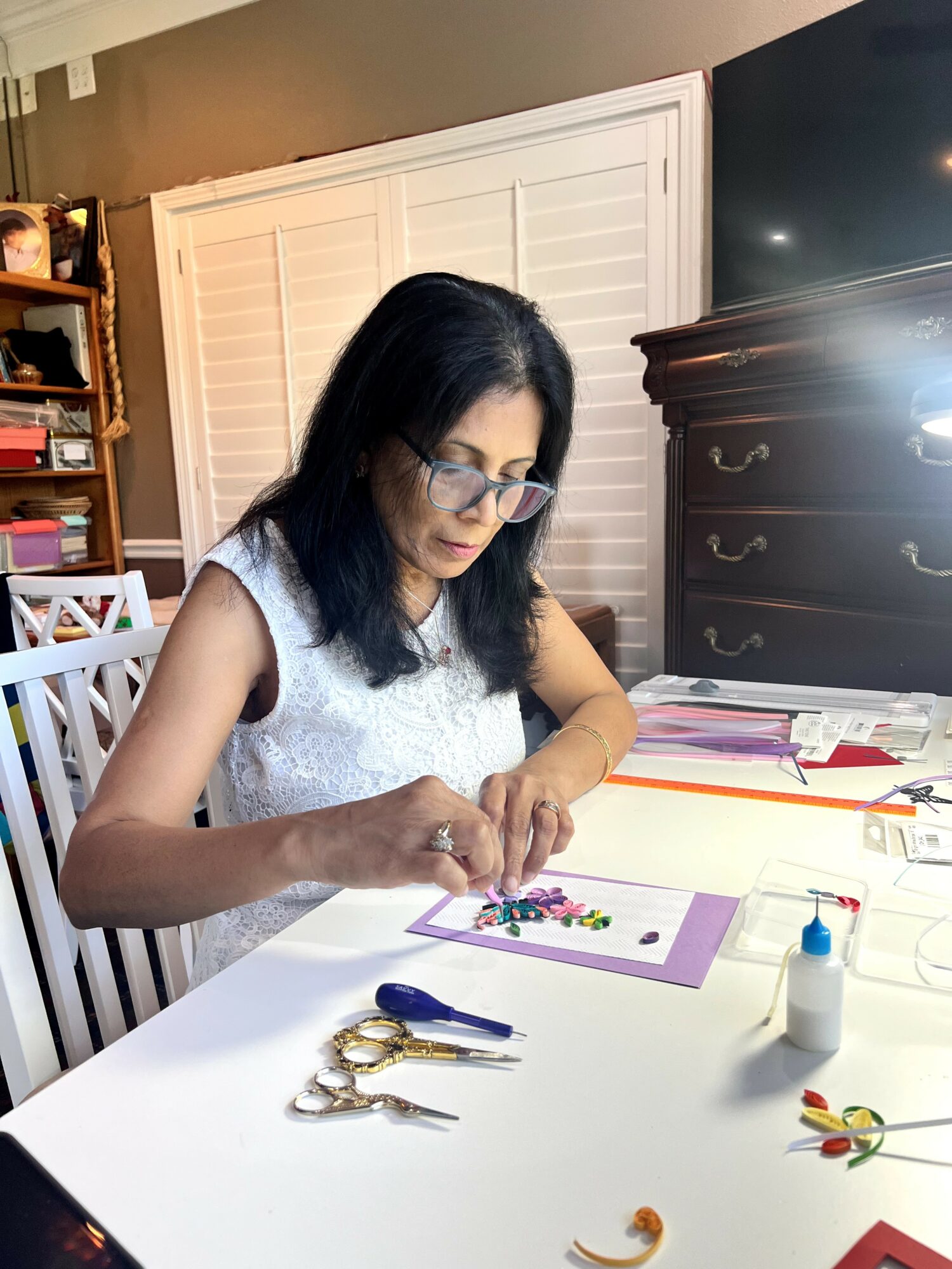 Woman with glasses working on a craft project at a table with scissors, glue, and paper, in a room with a dresser and bookshelf.
