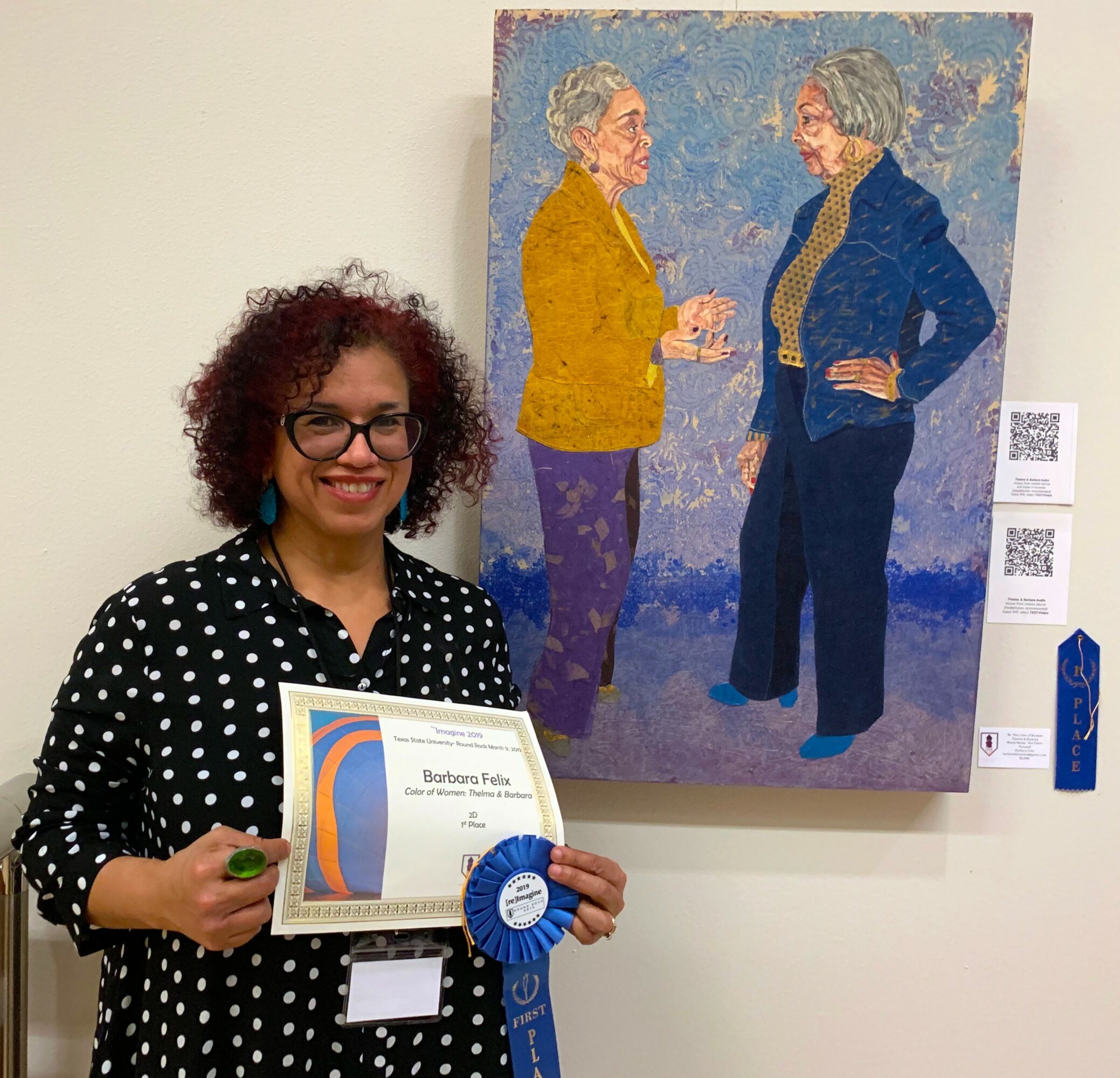 Woman with curly hair and glasses holding a certificate and blue ribbon, standing in front of a painting of two women talking.