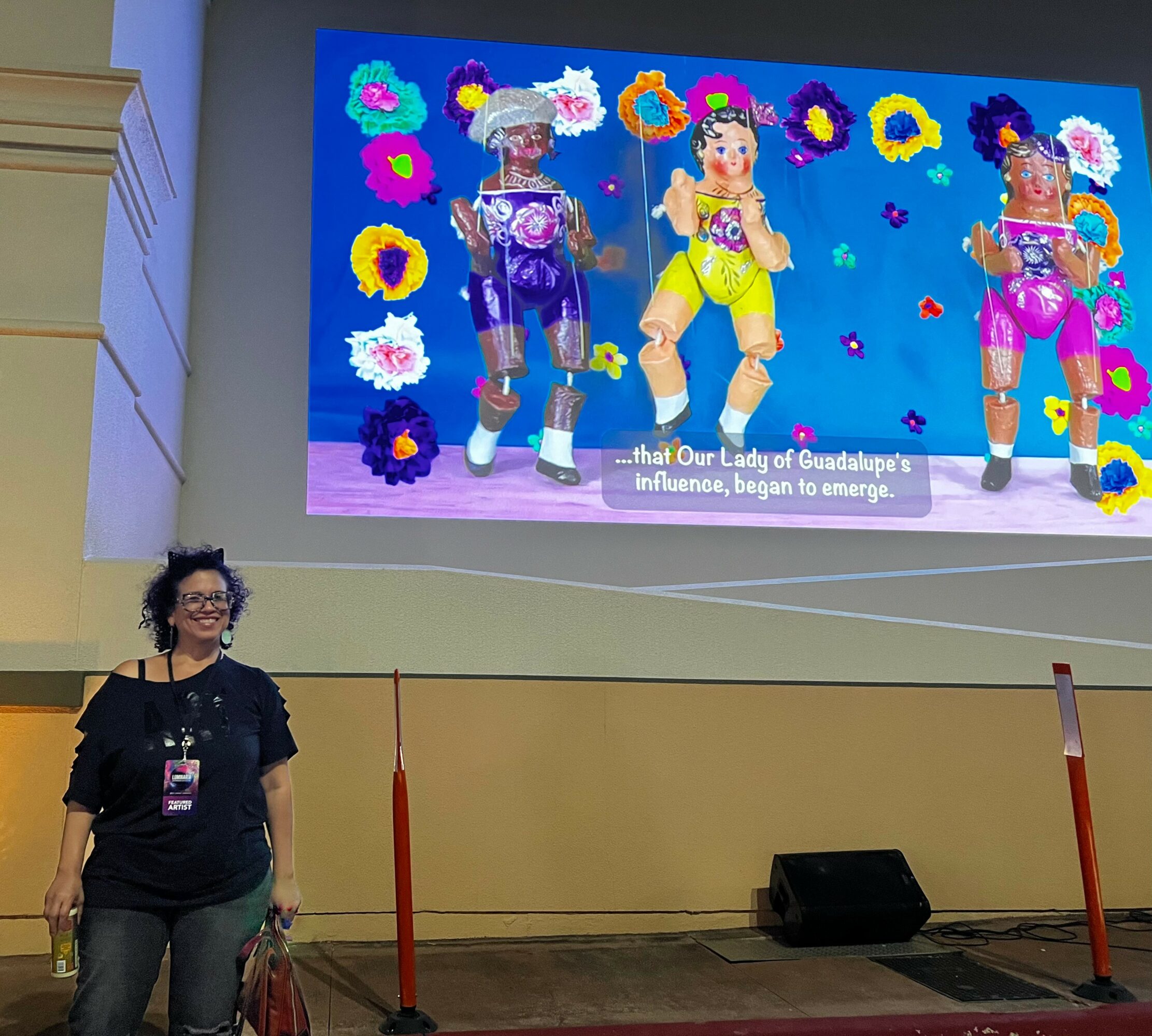 Woman standing in front of a large screen displaying colorful costumes and flowers, with a subtitle about Our Lady of Guadalupe's influence.