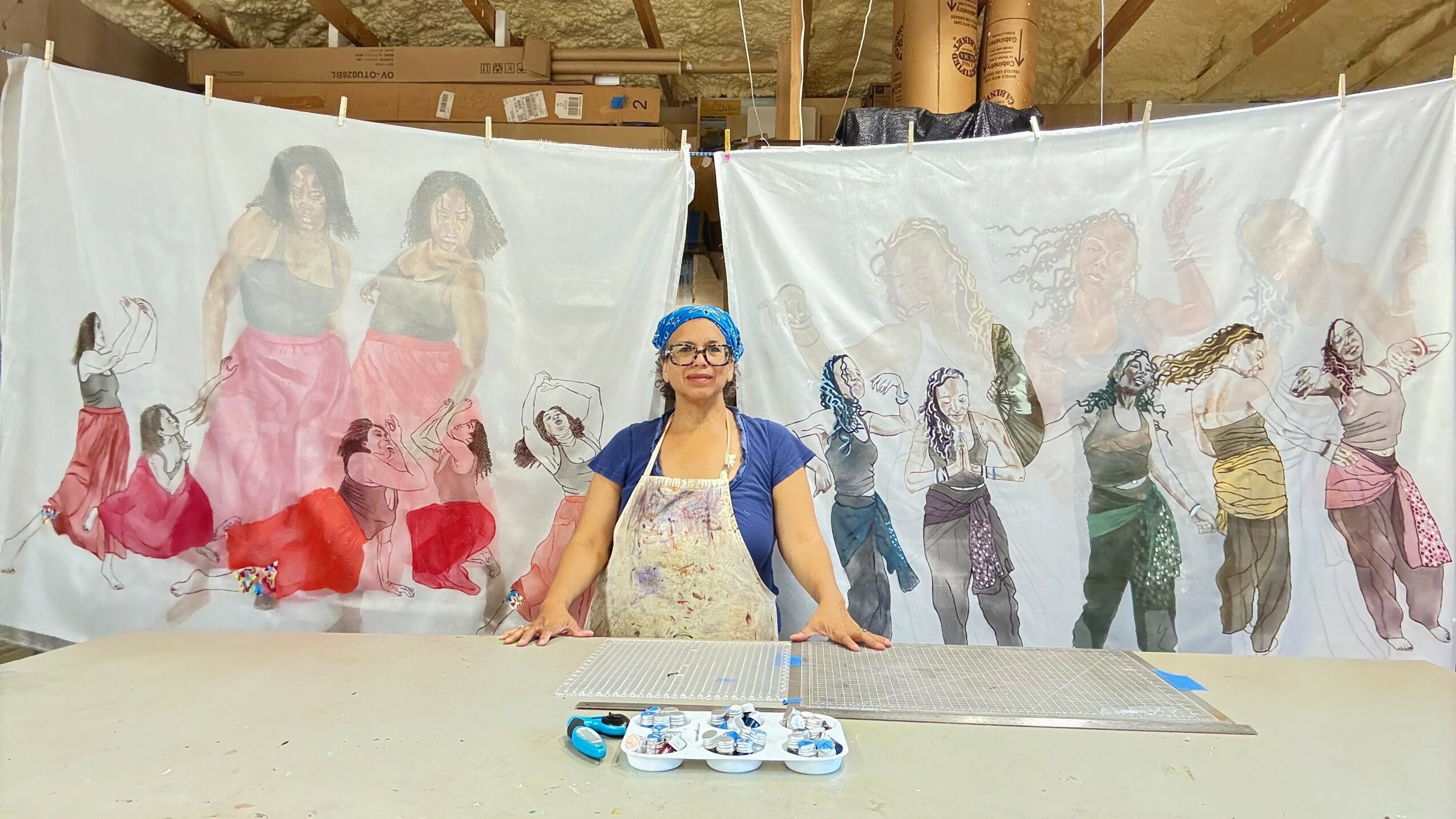 Woman standing in front of a backdrop with dancers in colorful costumes, warehouse setting, table with items in front.