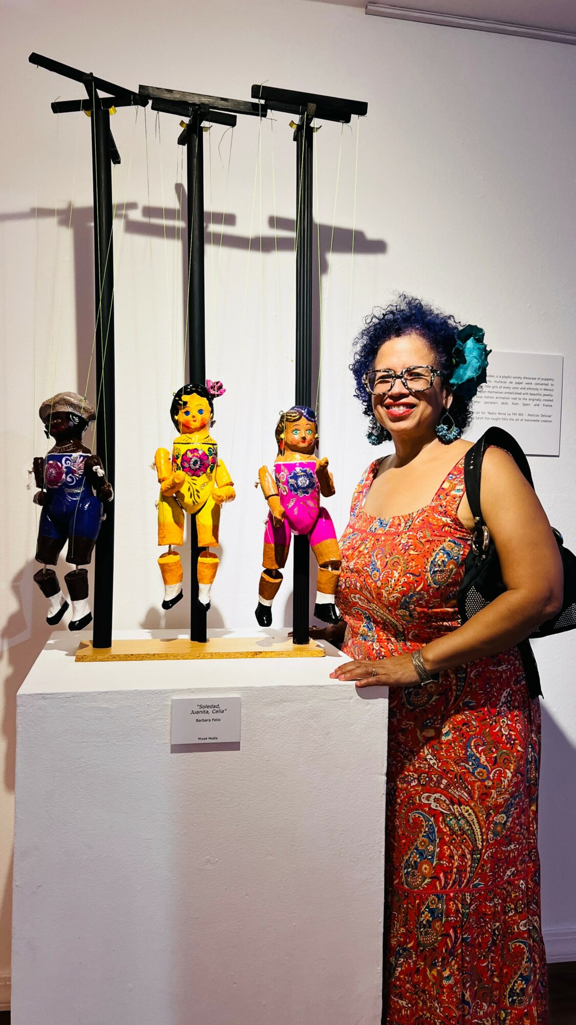 Woman with curly hair and glasses standing next to puppets on sticks, smiling, in an art gallery.