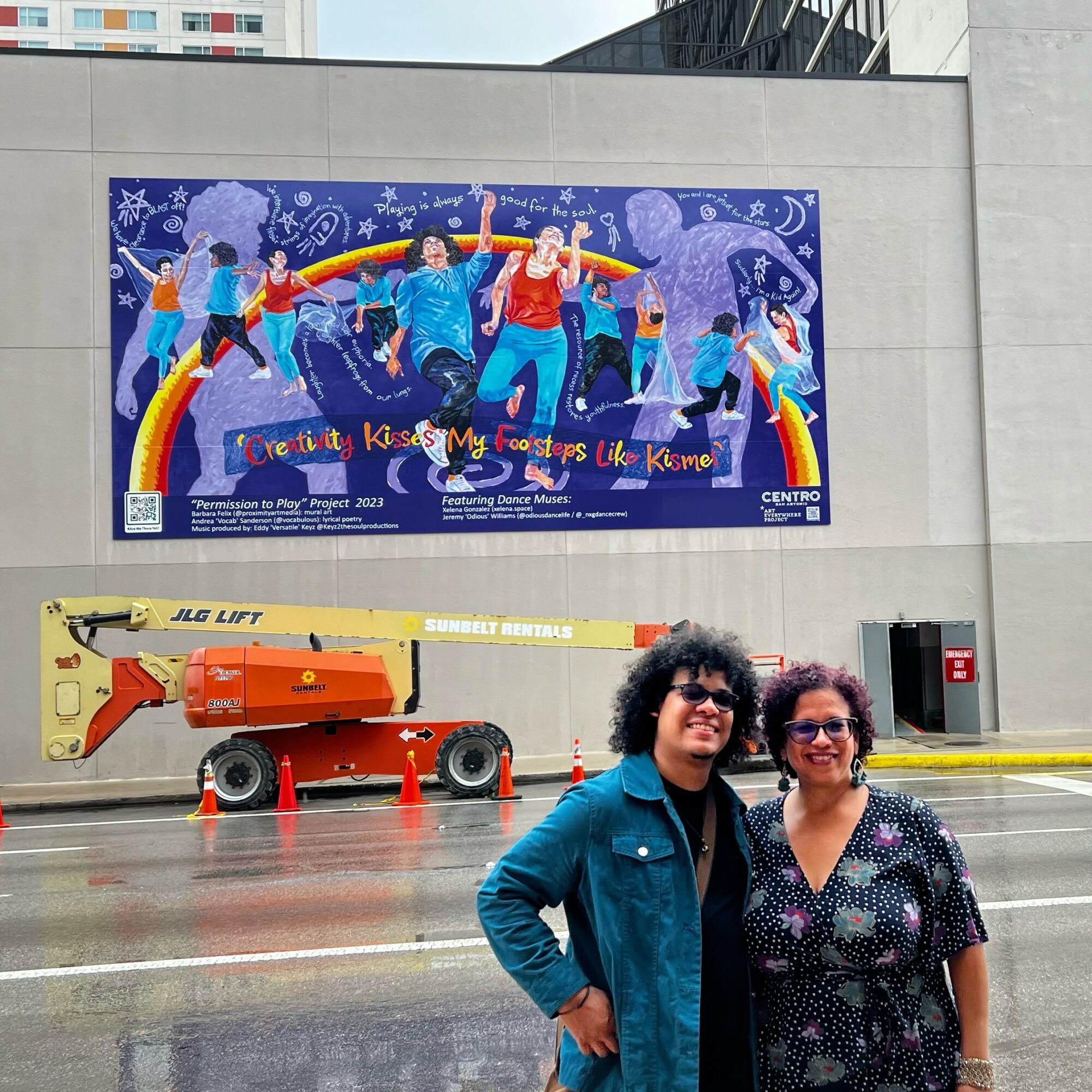 Two women stand in front of a mural and construction equipment on a city street, smiling at the camera.