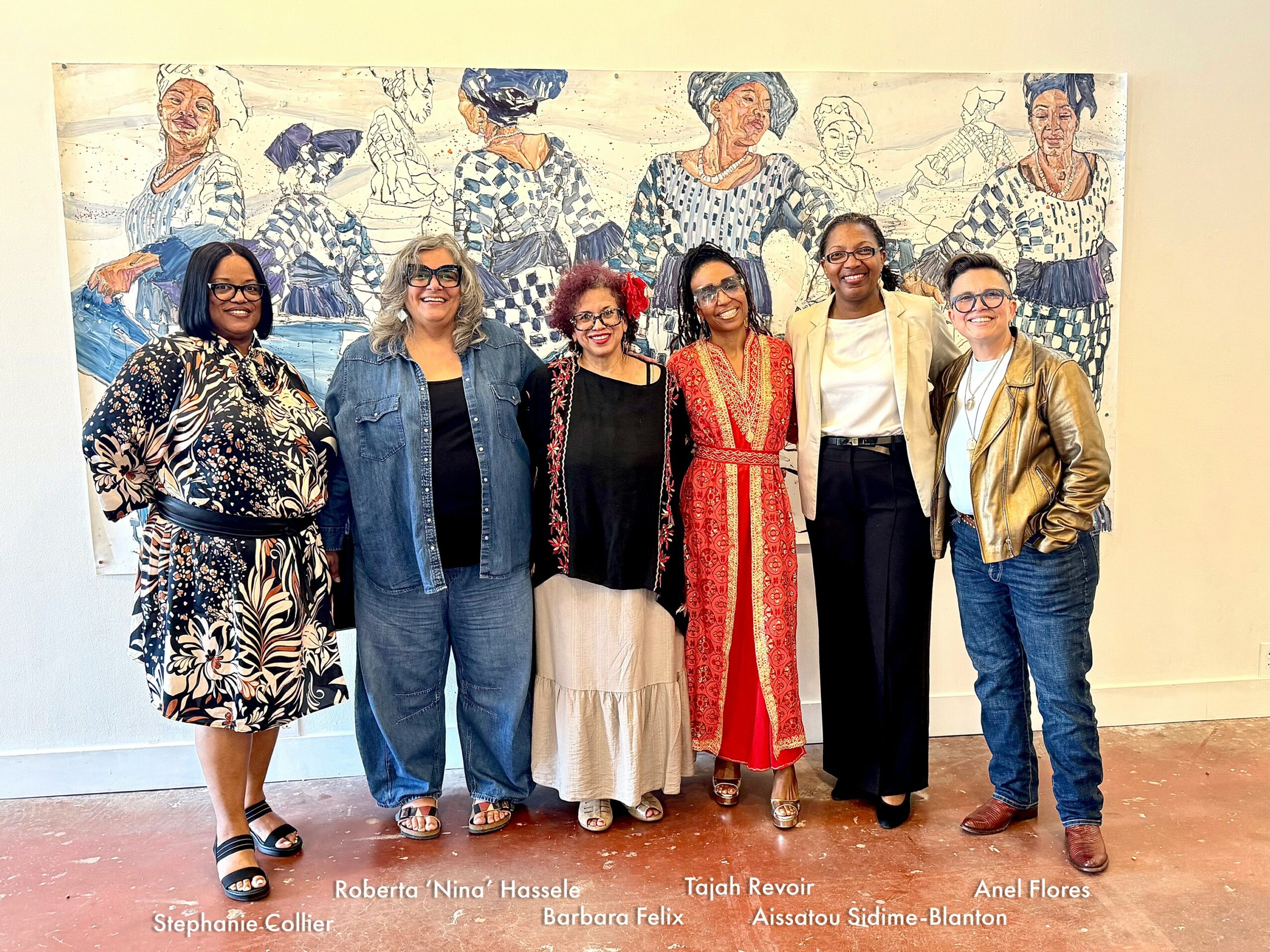 Group of six women standing in front of a mural with abstract figures, smiling, dressed in various outfits.