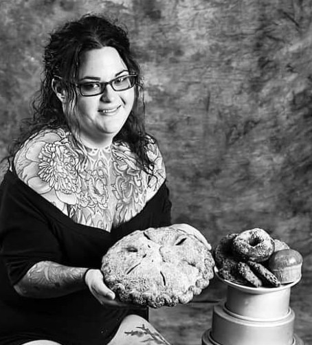 Woman with glasses holding a large pie, with a bowl of baked goods beside her, against a textured background.