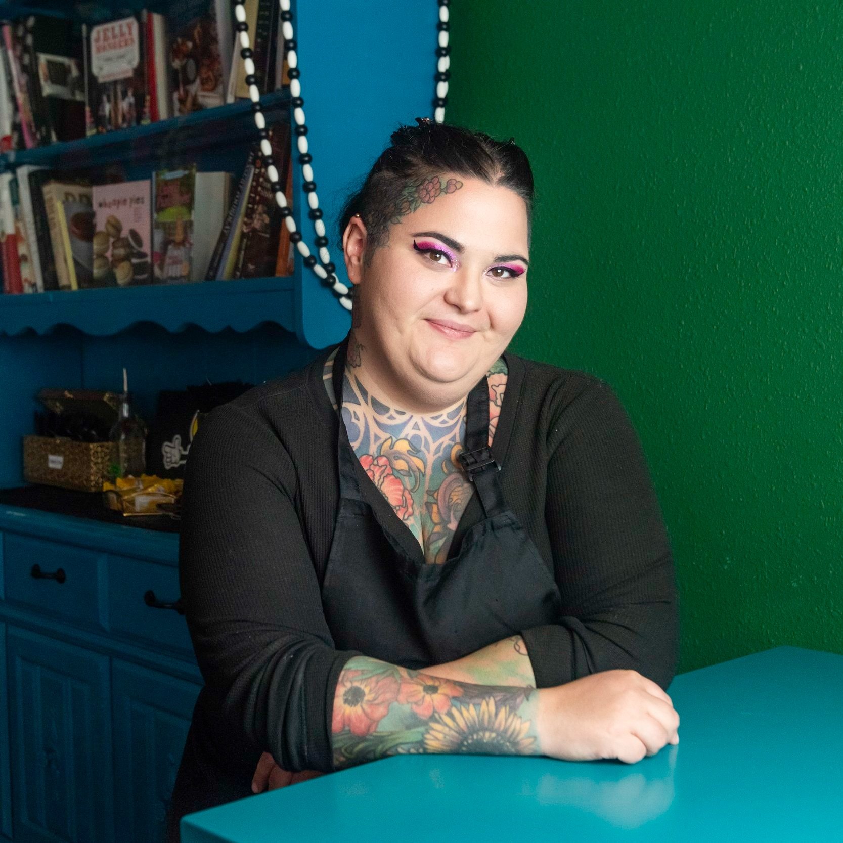 Person with dark hair, tattoos on arms and chest, smiling, sitting at a table in a colorful room with books and decorations.