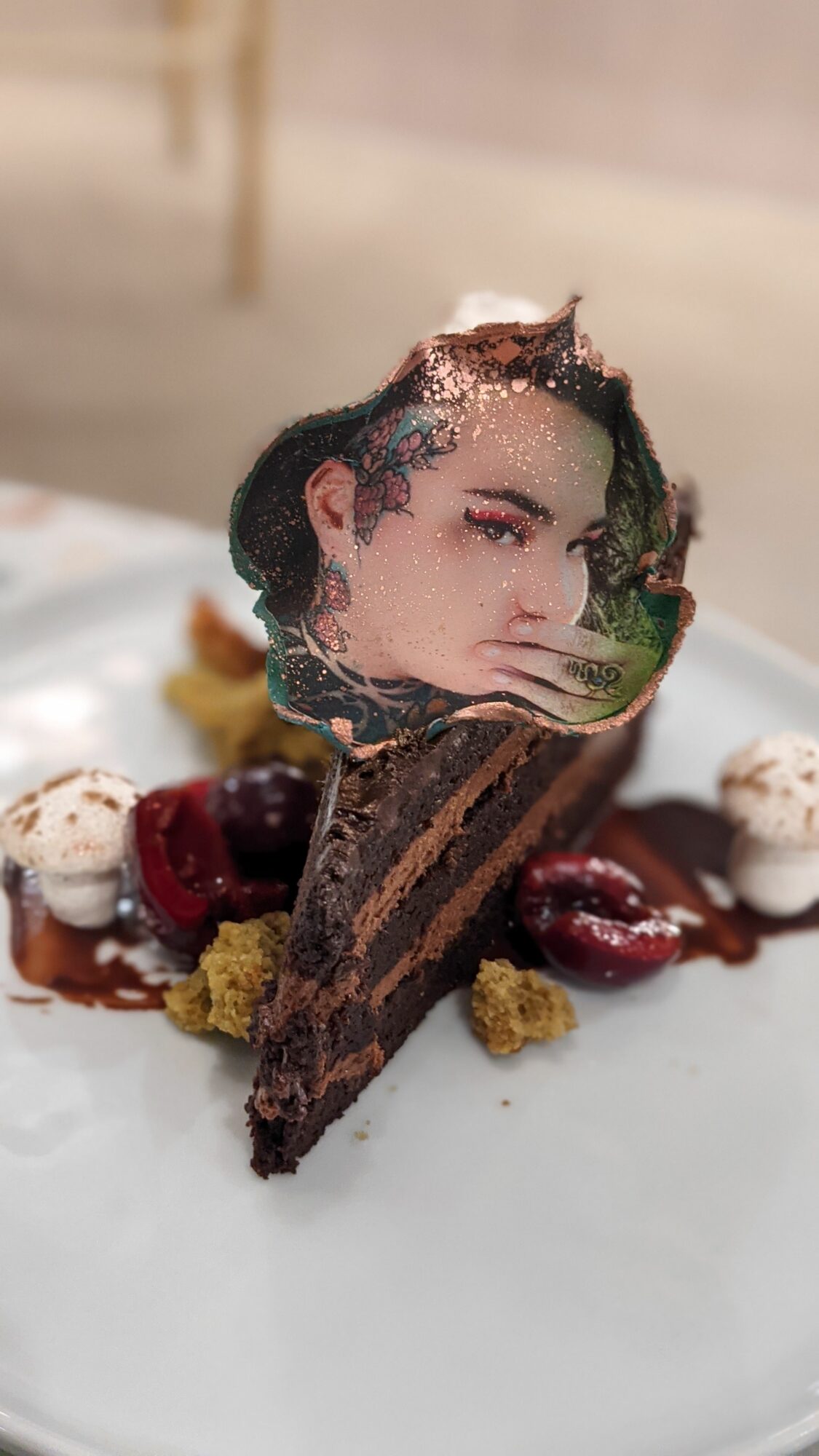 Dessert with a edible image of a woman's face, chocolate cake, cherries, and decorative toppings on a white plate.