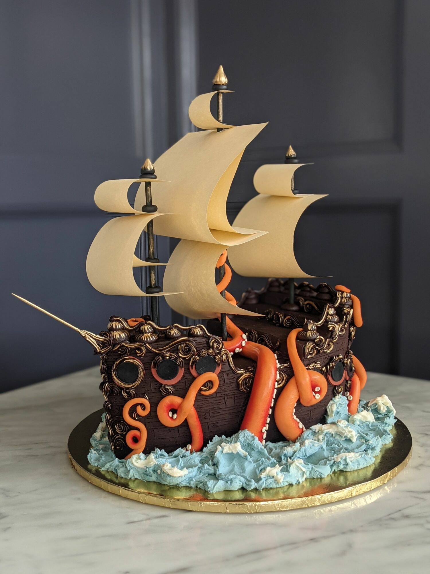 Decorative cake with tentacles and a ship sail, featuring blue and black icing, on a marble surface.