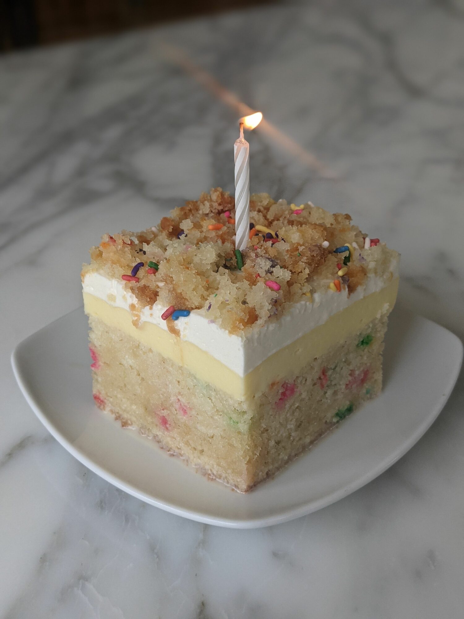 Slice of birthday cake with sprinkles and a lit candle on a white plate, on a marble surface.