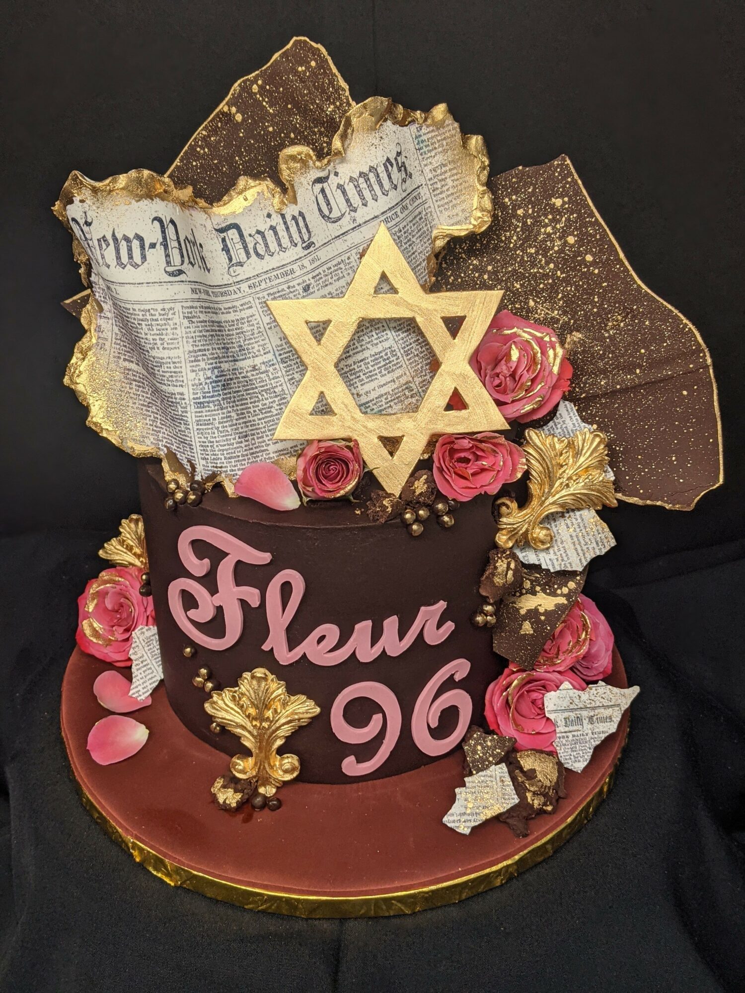 Chocolate cake with pink and gold decorations, including roses, a Star of David, and Hebrew text, on a black background.