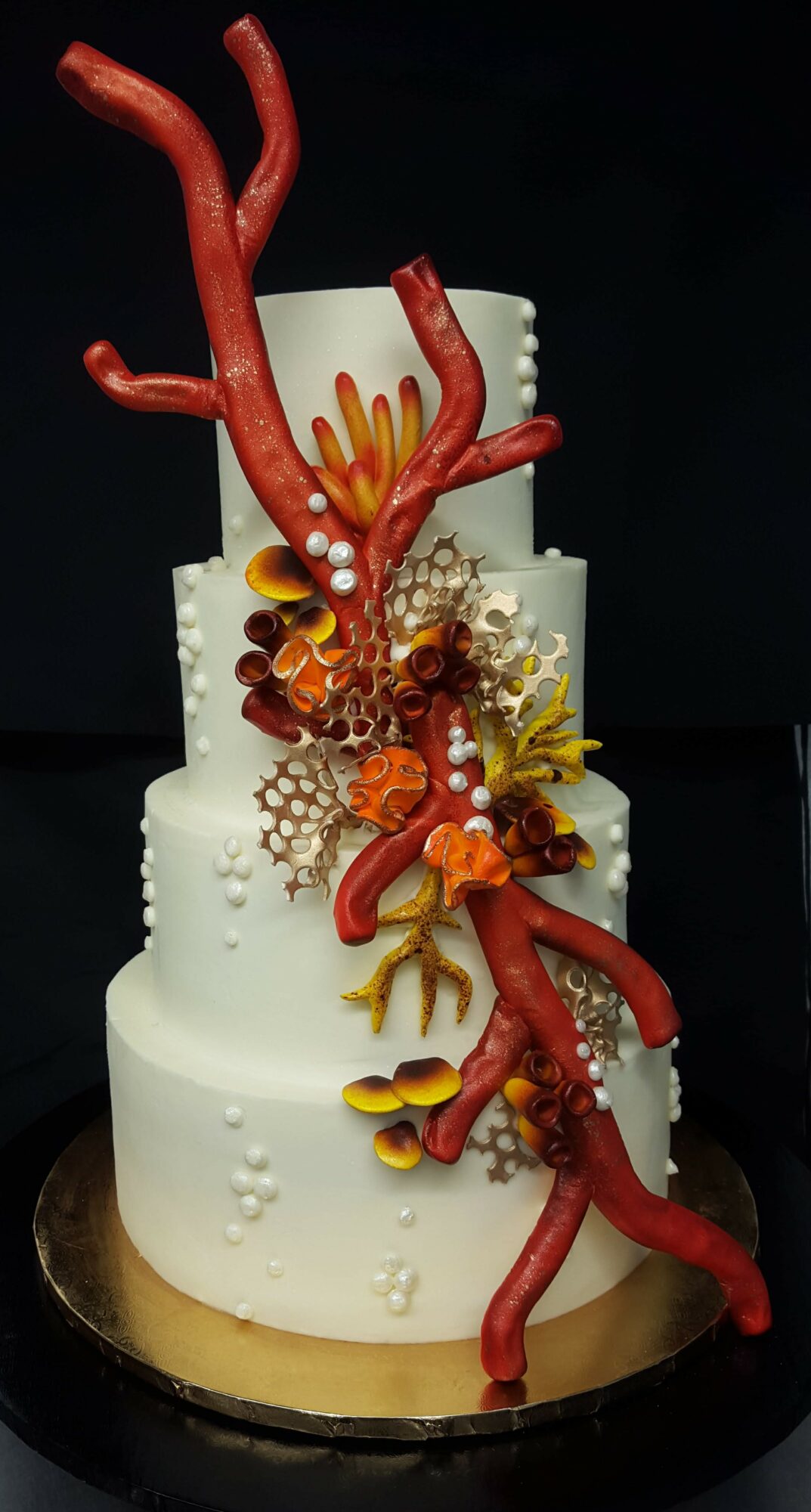 Three-tiered white cake decorated with red coral-like structures, gold accents, and small white flowers, on a black base.