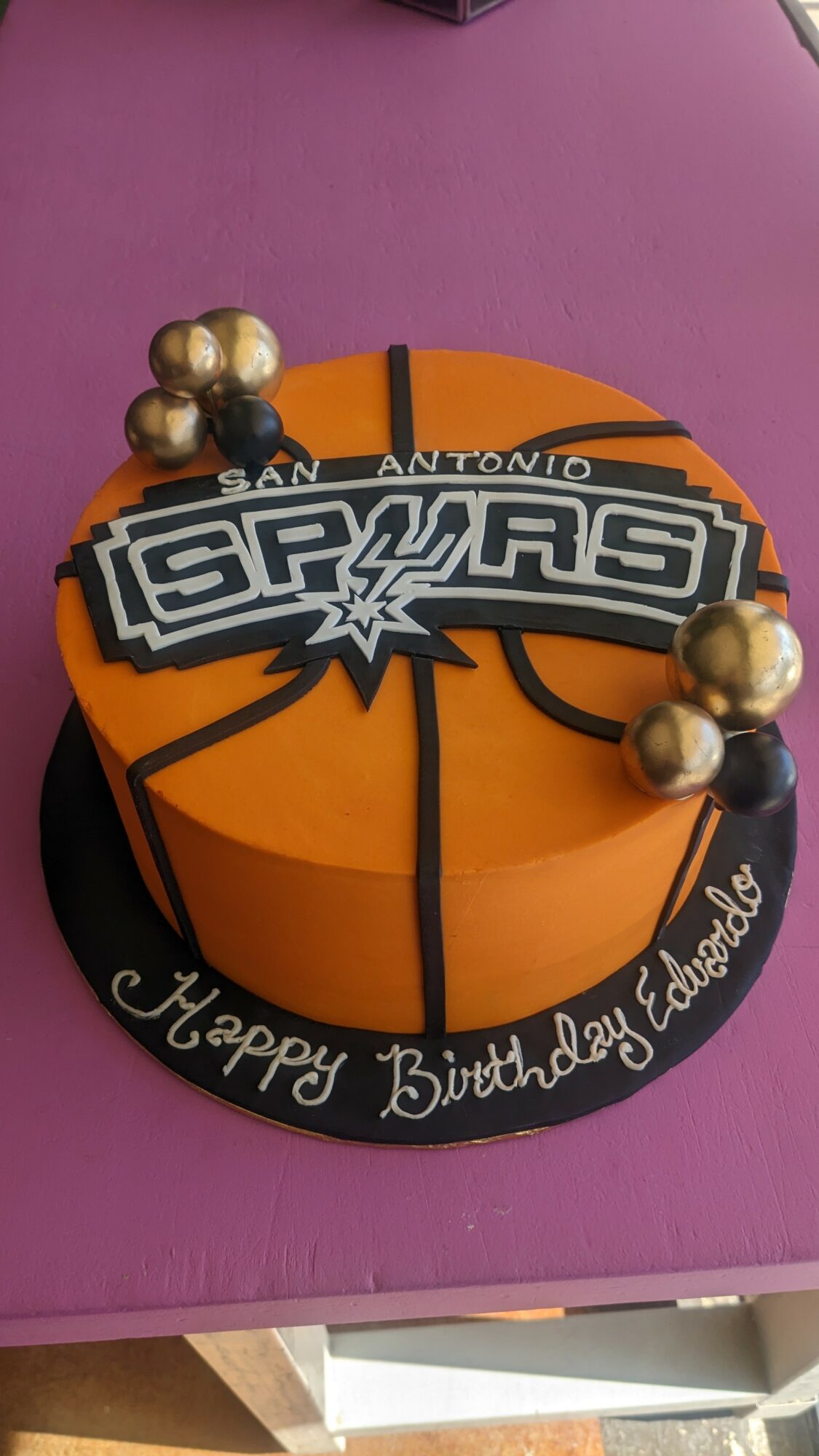 Round birthday cake decorated with a San Antonio Spurs logo, gold balls, and a black base with white writing, on a purple surface.