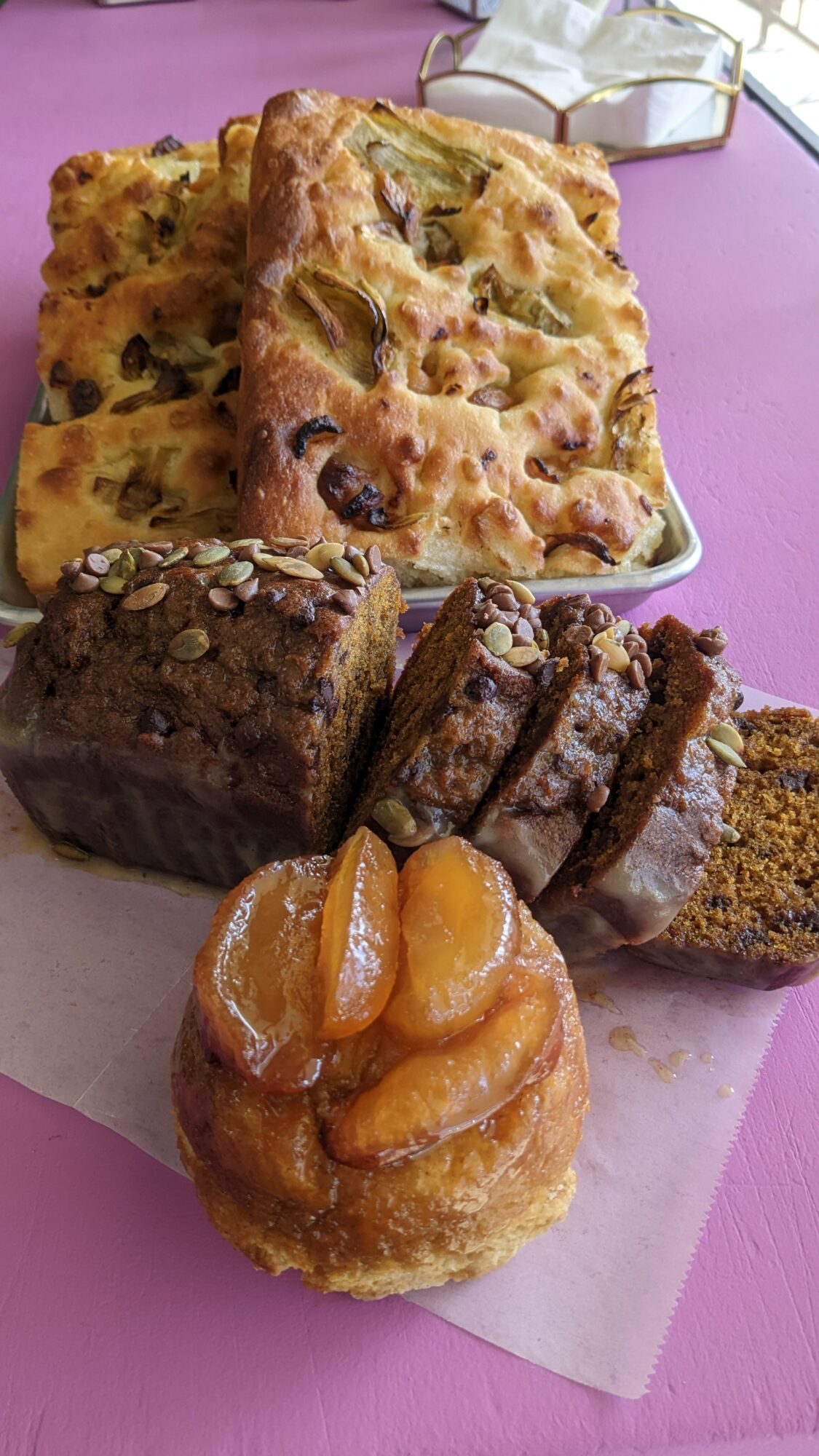 Assorted baked goods including bread, cake, and a pastry with fruit on top, on a pink surface.
