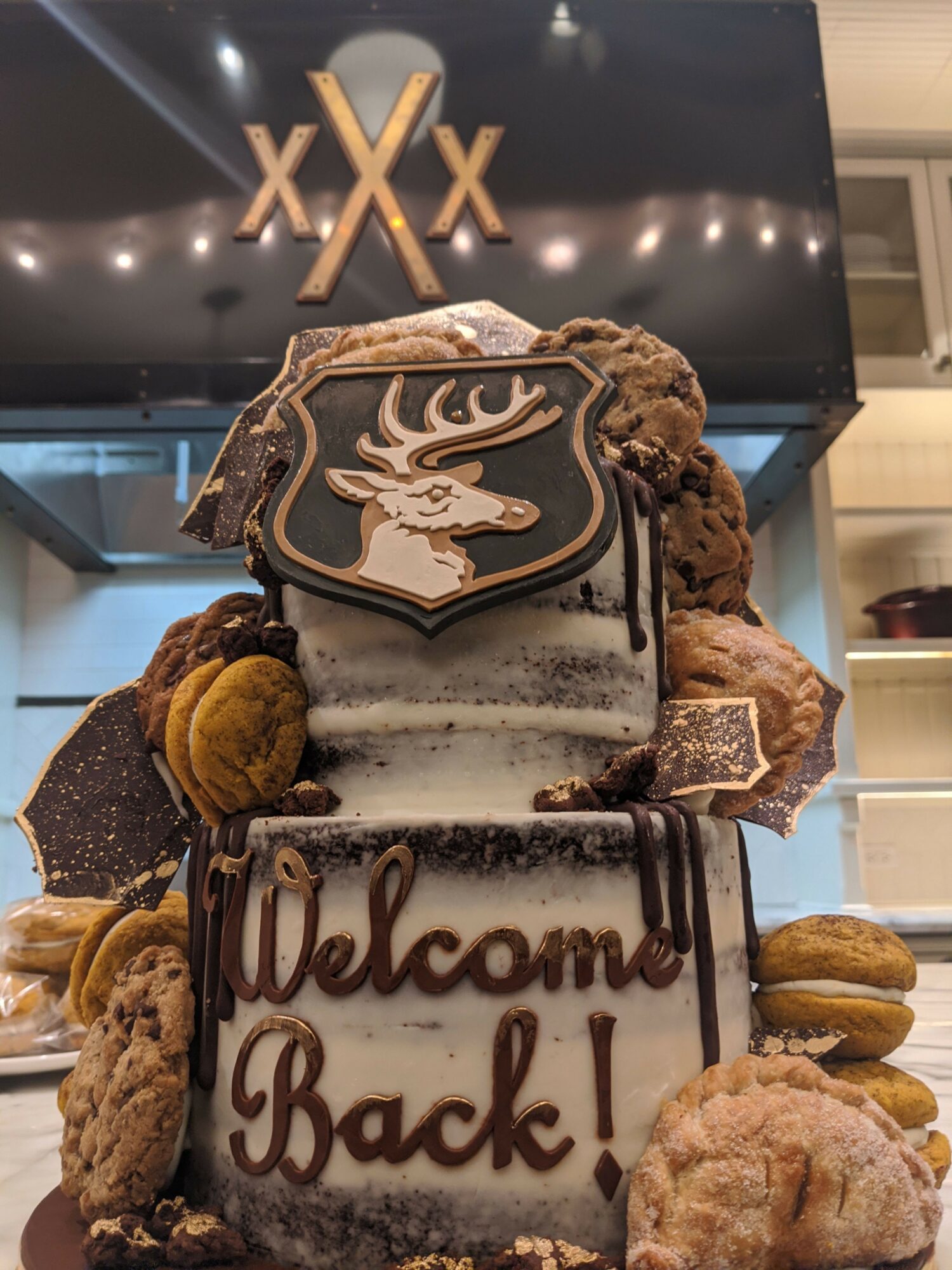 Two decorated cakes with cookies and chocolates on top and around, displayed on a table in a room with a black sign in the background.