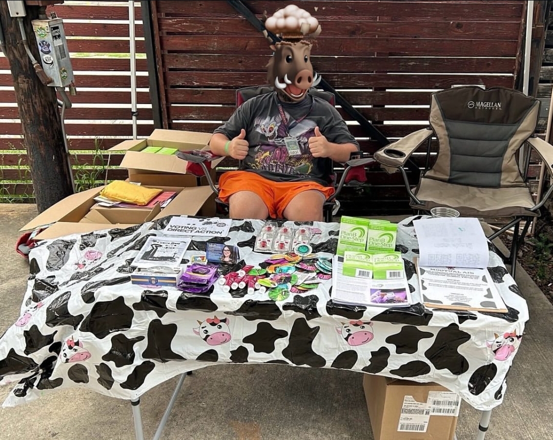Person wearing a horse mask sitting behind a table with various items, outdoors with a wooden fence and chairs, cow-patterned tablecloth.