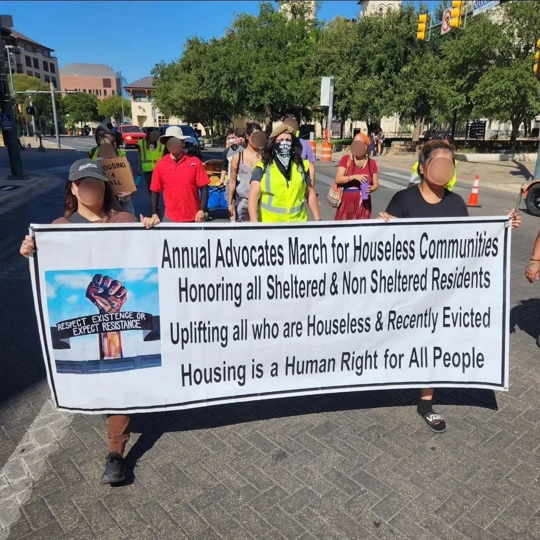 Group of people holding a banner at a march, with trees and buildings in the background, sunny day.