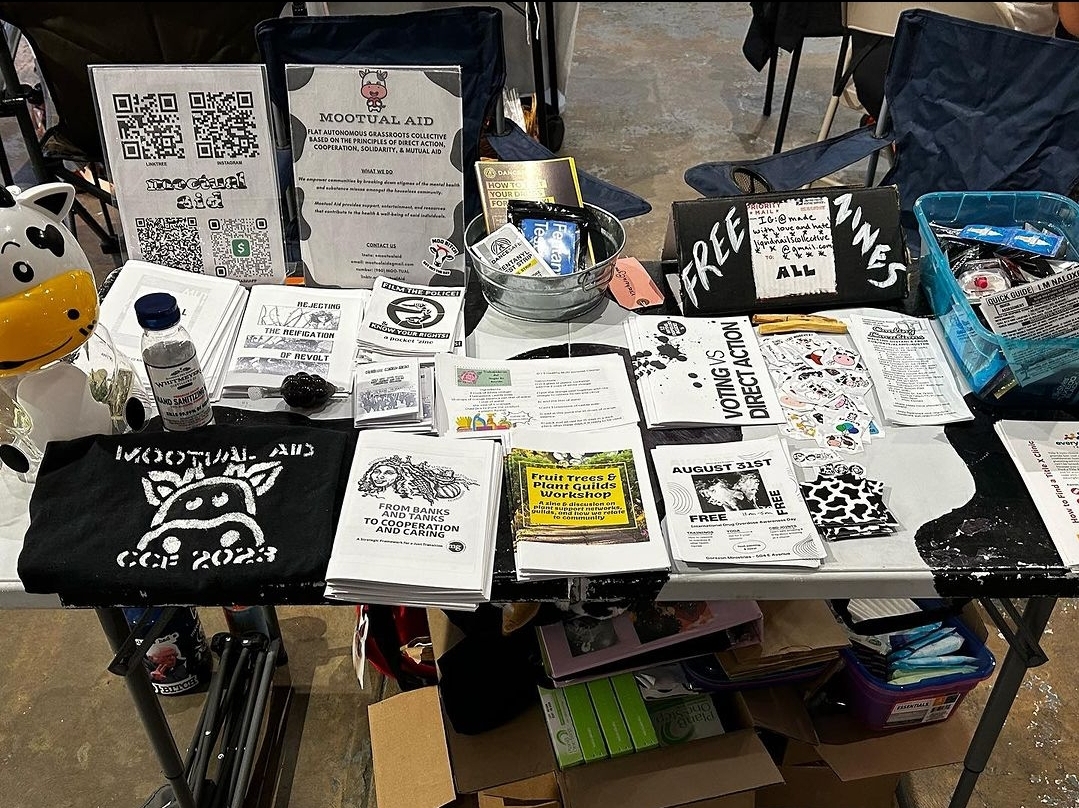 Table with informational materials, a stuffed animal, and various supplies, set outdoors with chairs around it.