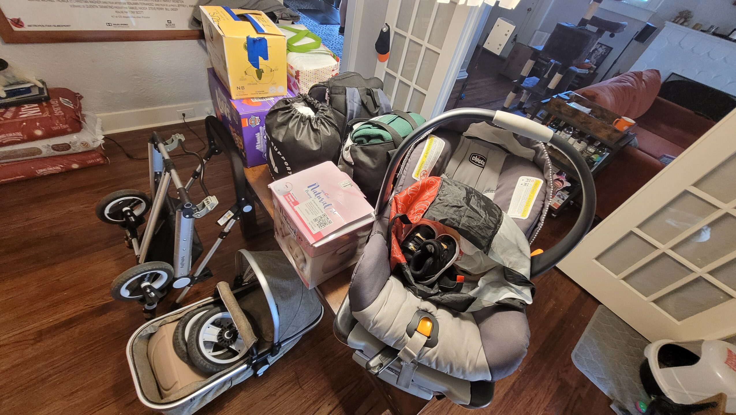 Baby car seat, stroller, boxes, and various items on a wooden table, with a window and room in the background.