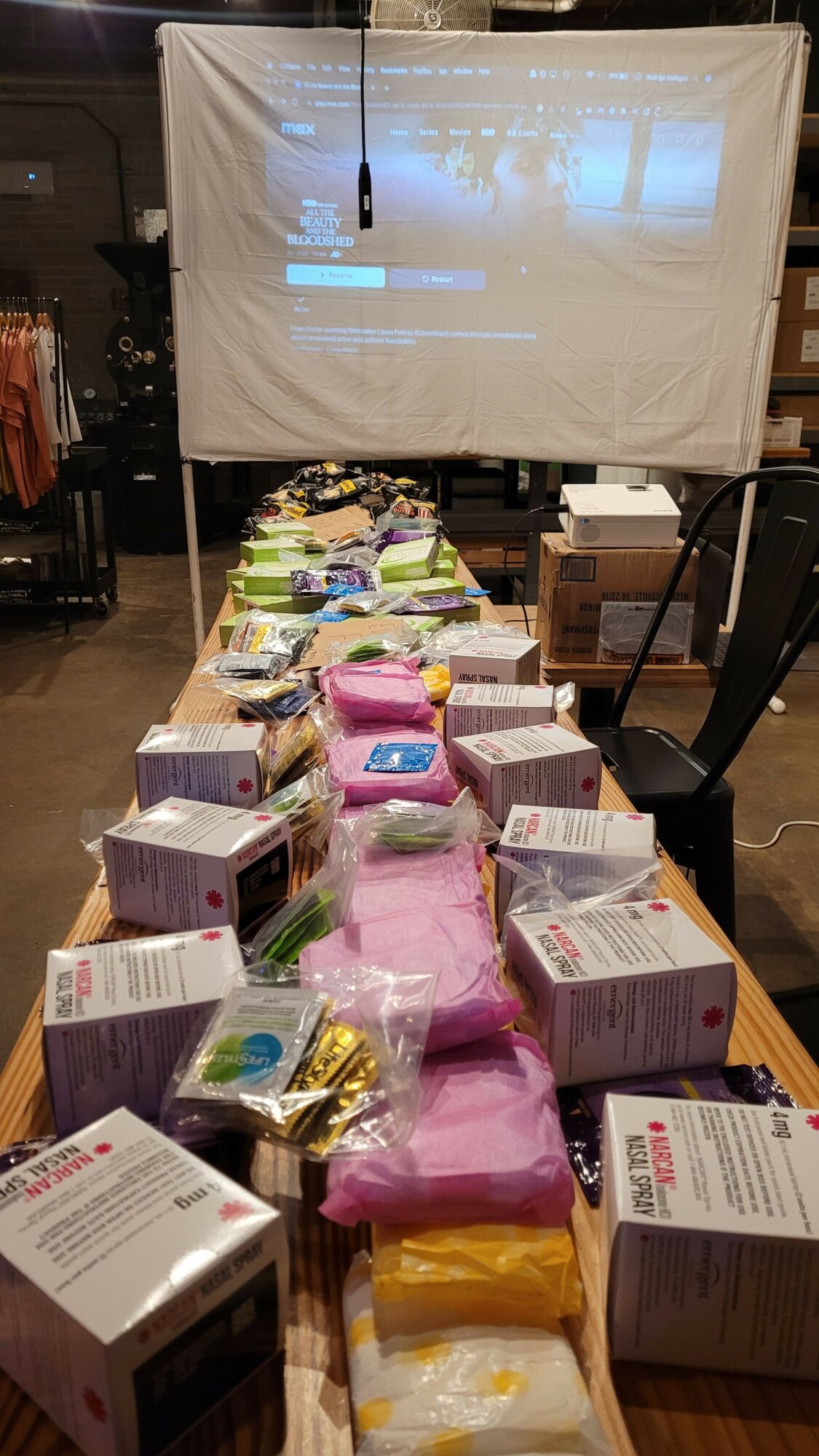 Table with various boxed products and pink packaging, set in a room with a projection screen in the background.