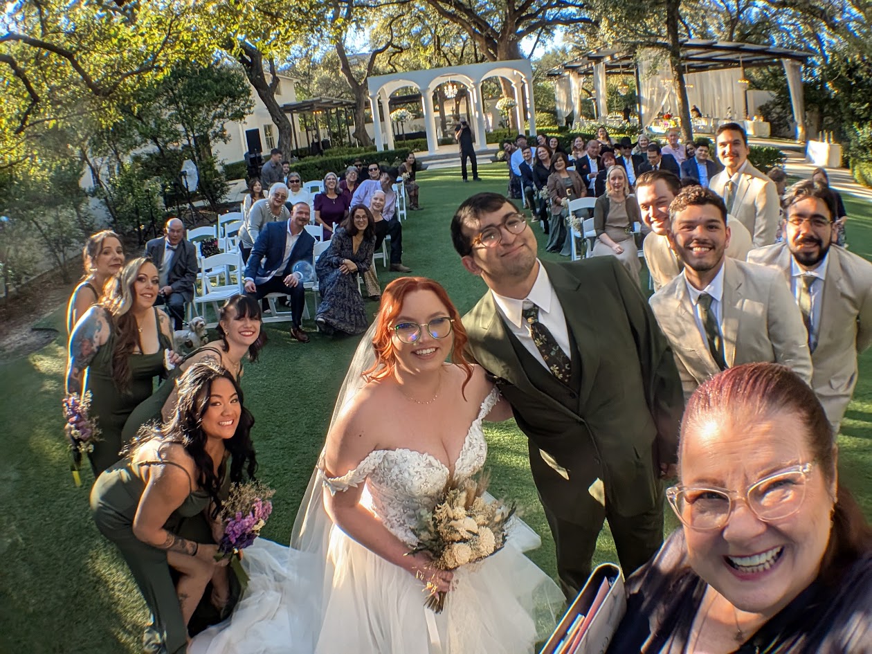 Wedding guests gathered outdoors, smiling, with a bride holding a bouquet, surrounded by friends and family, trees, and decorative arches.