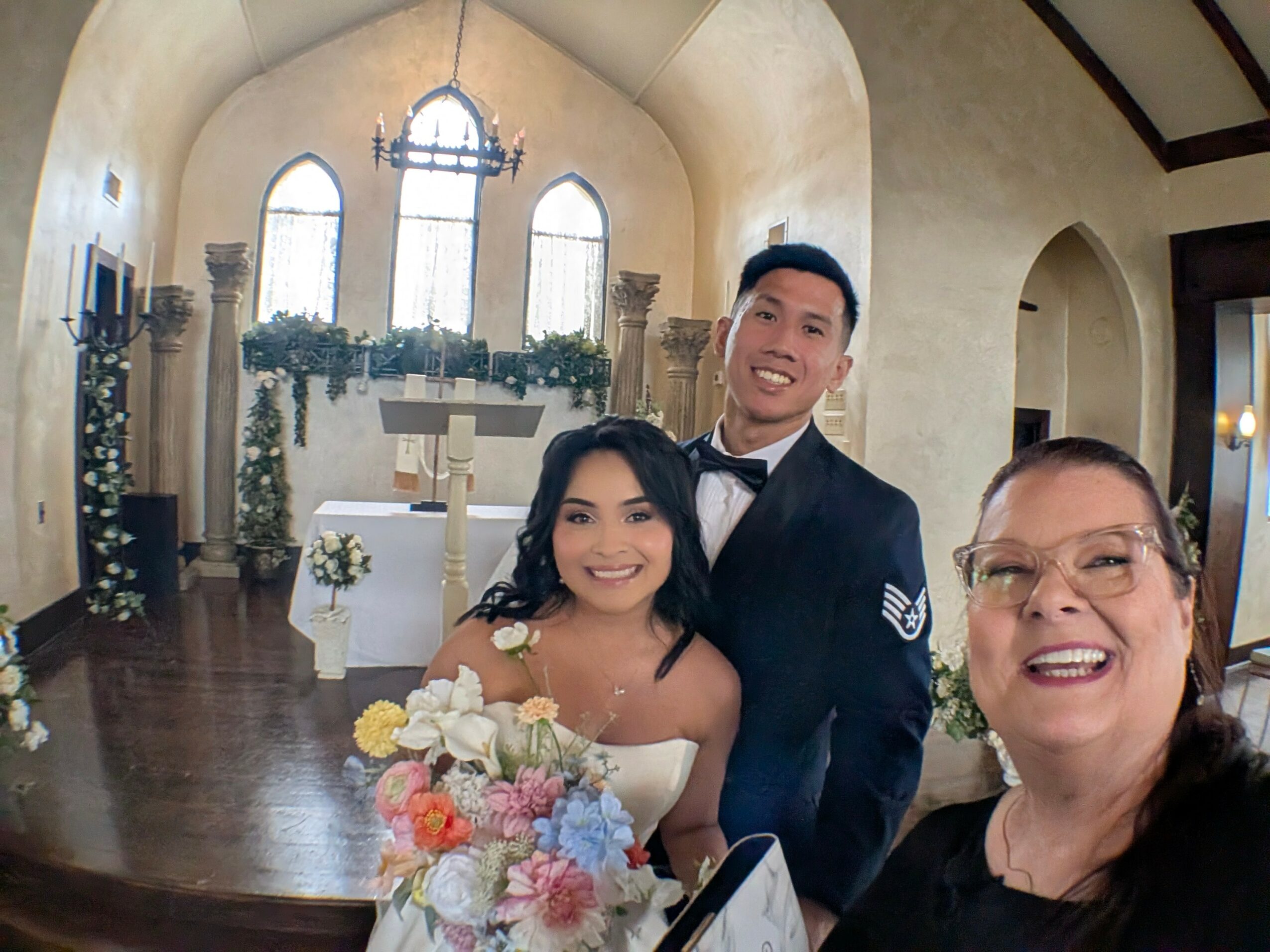 Three people smiling inside a church, with a woman holding a bouquet, a man in a suit, and another woman wearing glasses.