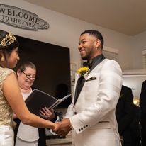 A man and woman hold hands during a wedding ceremony, with a woman reading from a book in the background.