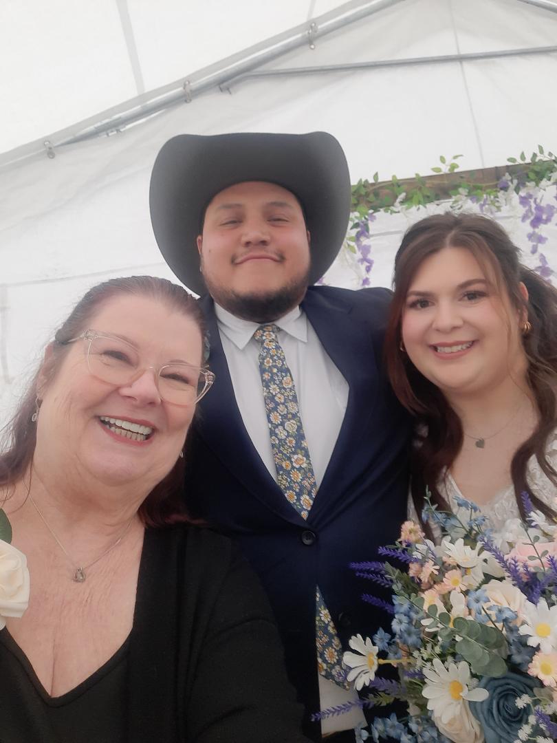 Three people smiling, one wearing a cowboy hat, holding flowers, in a bright indoor setting.