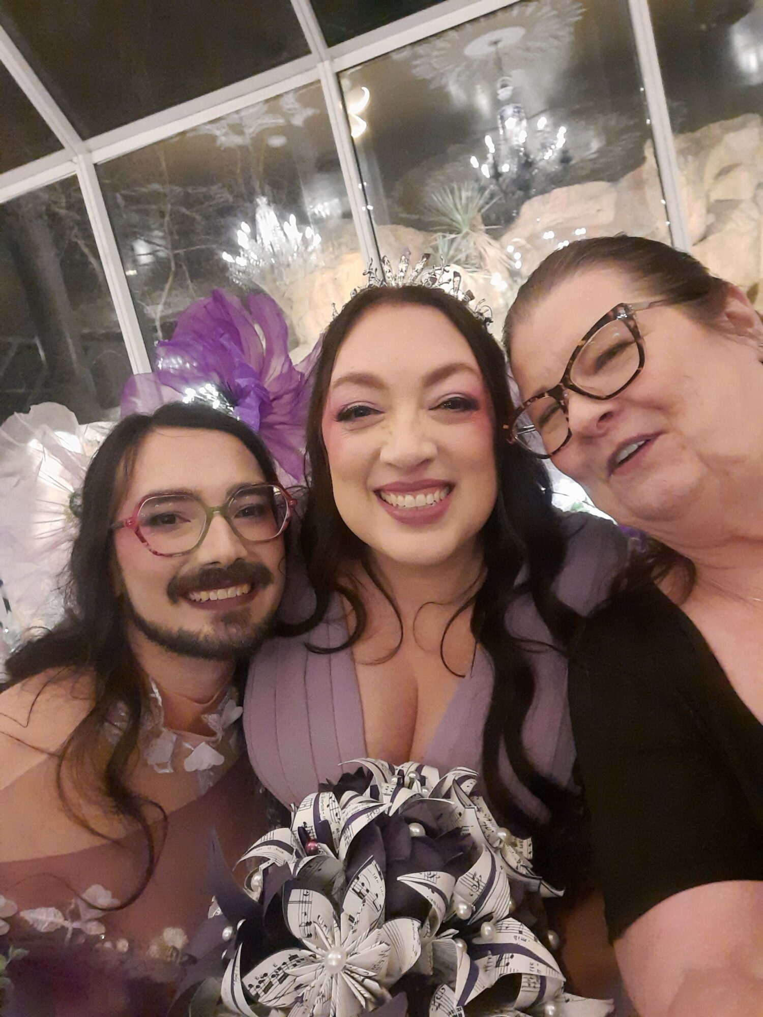 Three people smiling and taking a selfie indoors with a window and lights in the background.