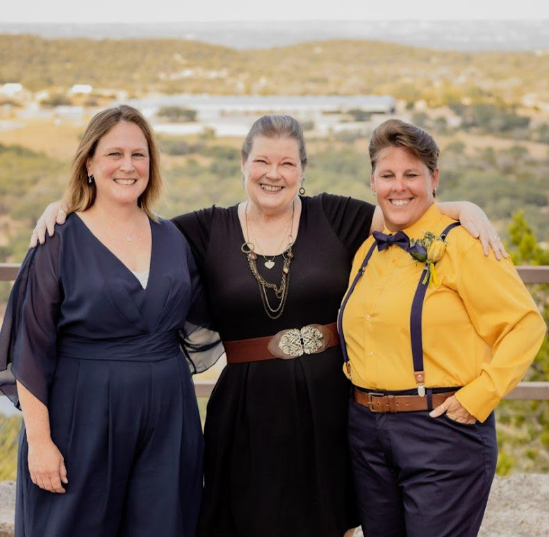 Three women standing outdoors, smiling, with a landscape background, arms around each other.