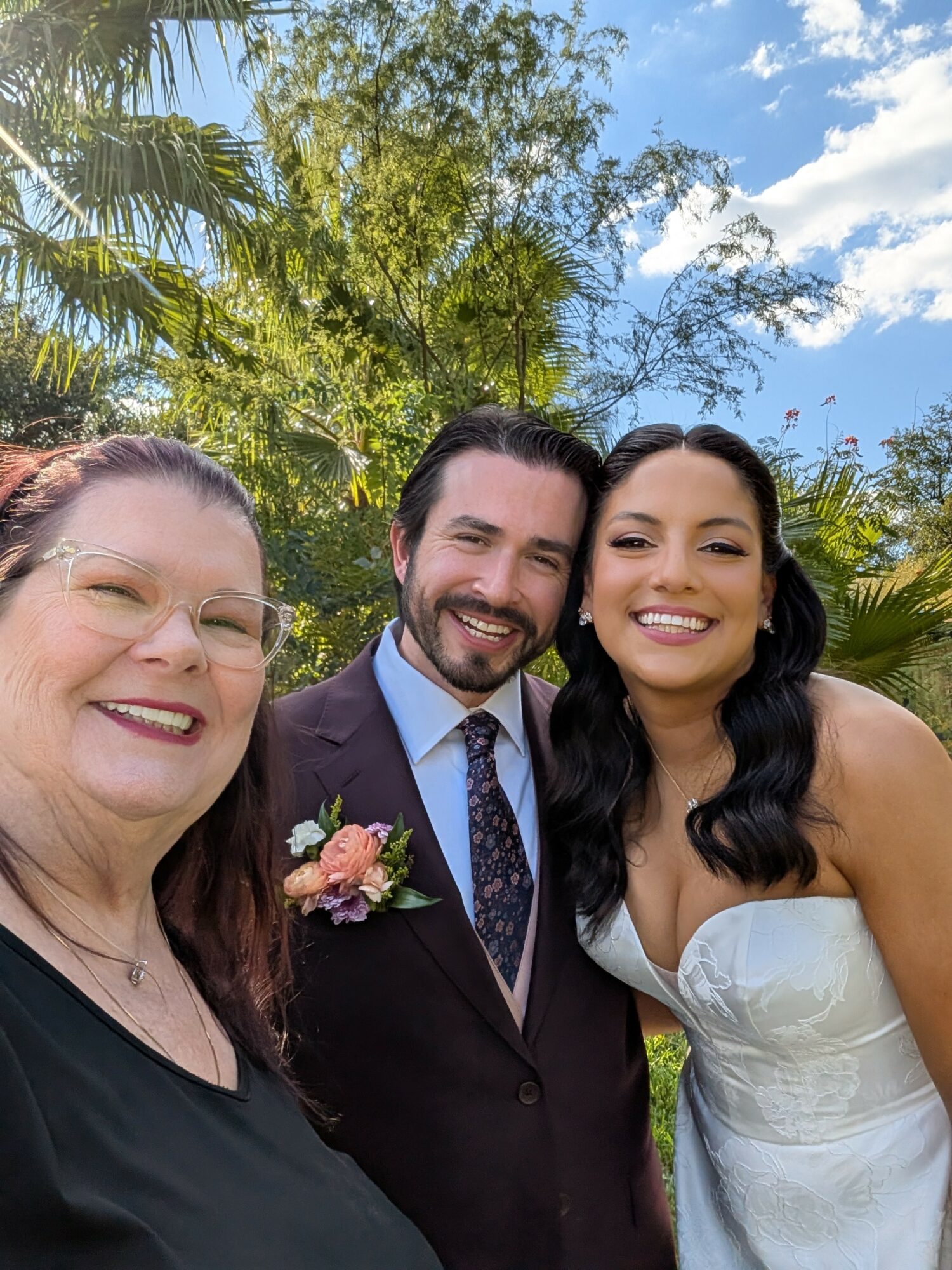 Three people smiling outdoors with trees and blue sky in background.