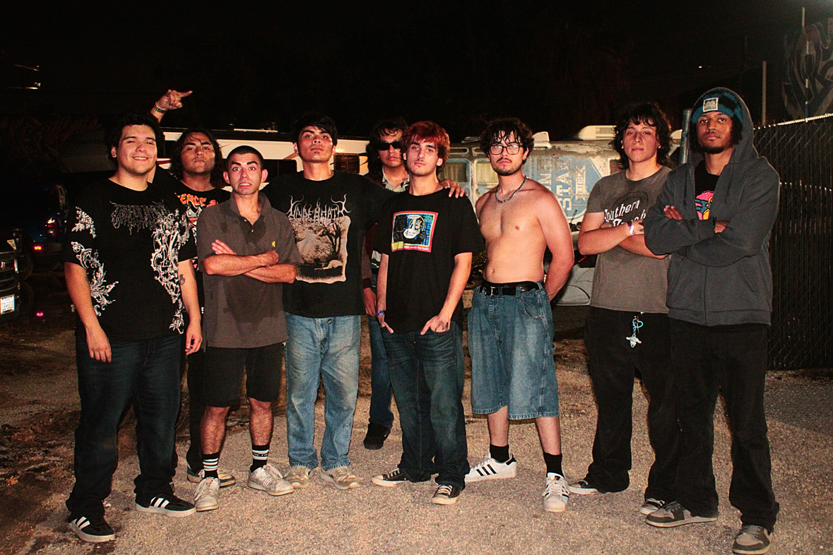 Group of nine young men standing outdoors at night, some with crossed arms, one shirtless, in casual clothing.