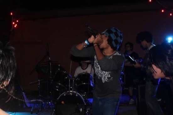 Person singing into a microphone on stage with band members in background, dim lighting, and colorful lights.