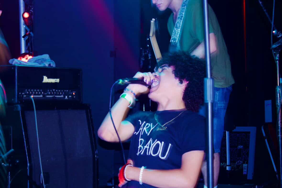 Person singing into a microphone on stage with musical equipment, another person playing guitar in the background, dark stage lighting.