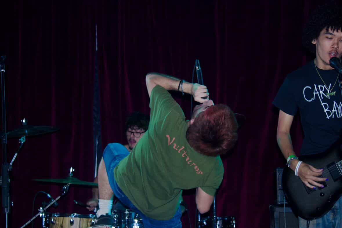 Person with short hair in a green shirt holding a microphone on stage with a band, dark red curtain background.