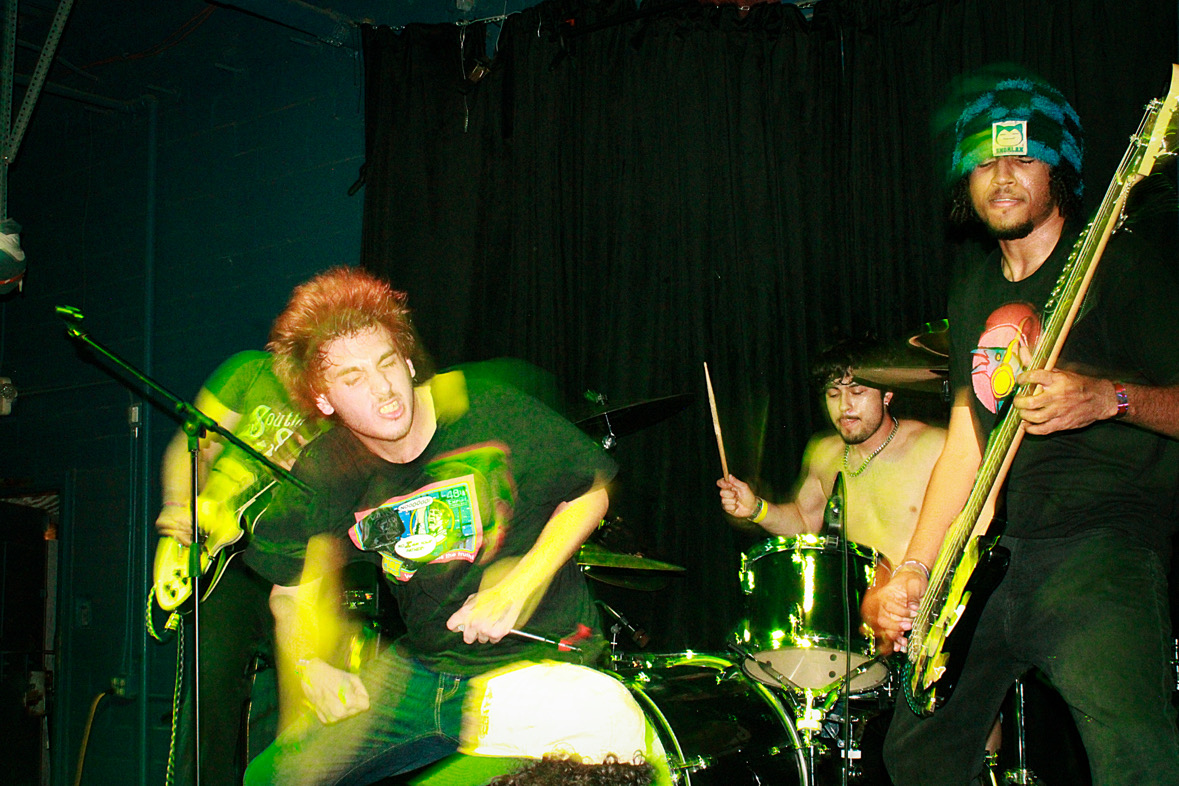 Three musicians performing on stage with drums and bass guitar, dark background, one person with curly hair, another with a cap.