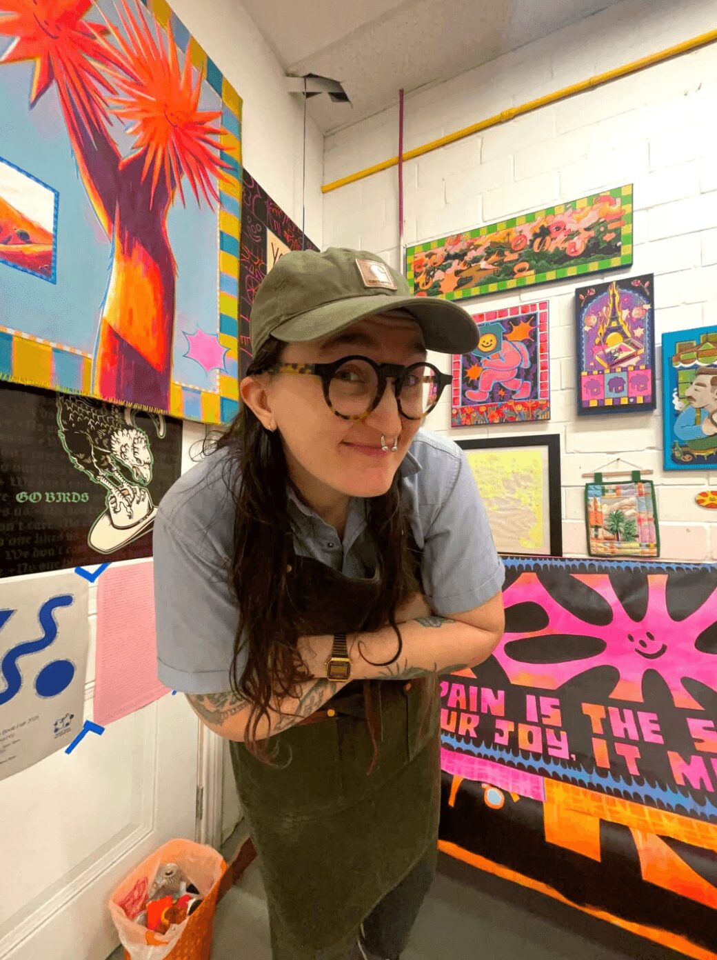 Person with long dark hair, glasses, and a cap, standing in front of colorful artwork on gallery wall.