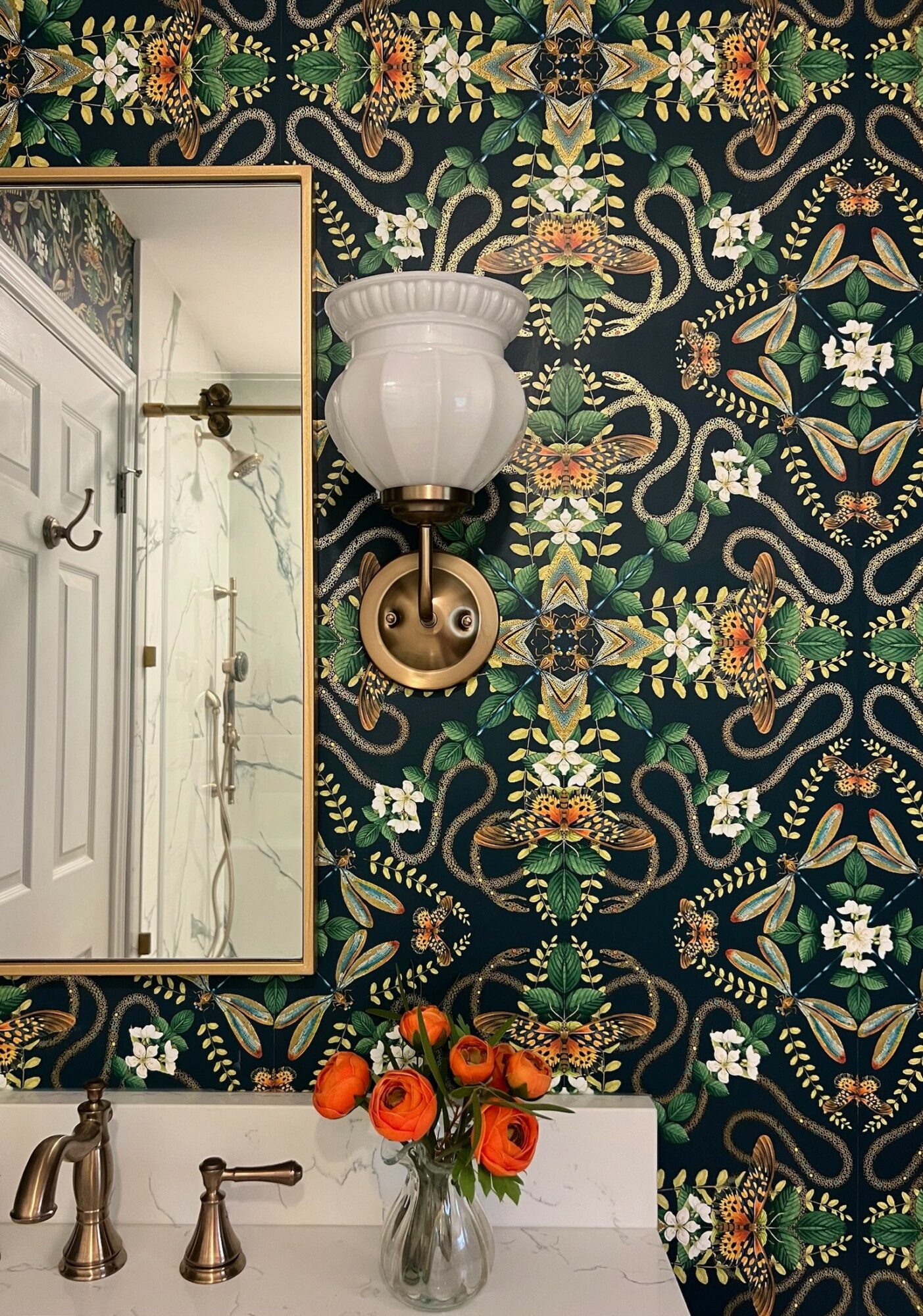 Wall-mounted light fixture with a white glass shade above a mirror, floral wallpaper background, and a vase of orange flowers.